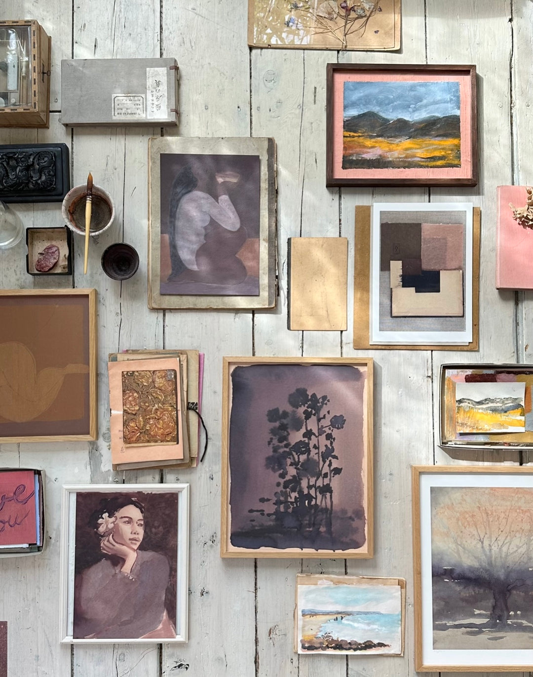 A wall display featuring multiple framed artworks, including the same painting of the woman holding a bowl, a portrait of a woman with a flower in her hair, a landscape, and various abstract and botanical pieces arranged in a gallery style.