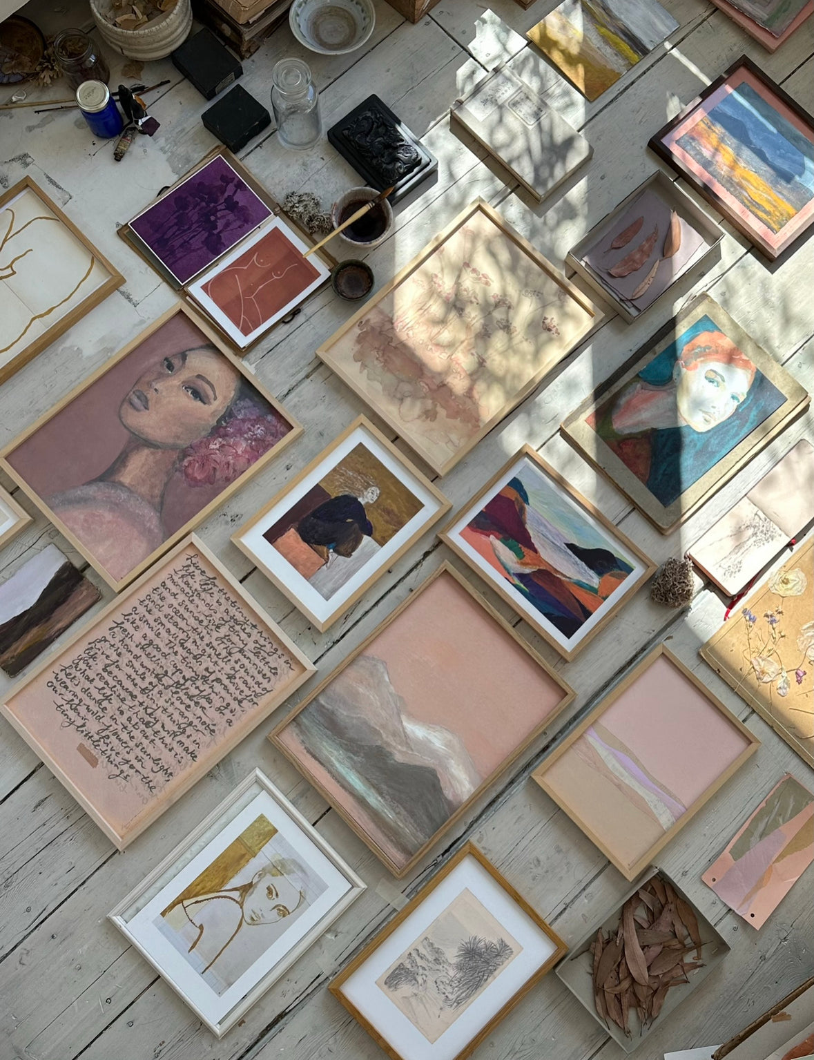 Collection of framed abstract, figurative, and botanical artworks including handwritten poetic text laid out on rustic wooden floor