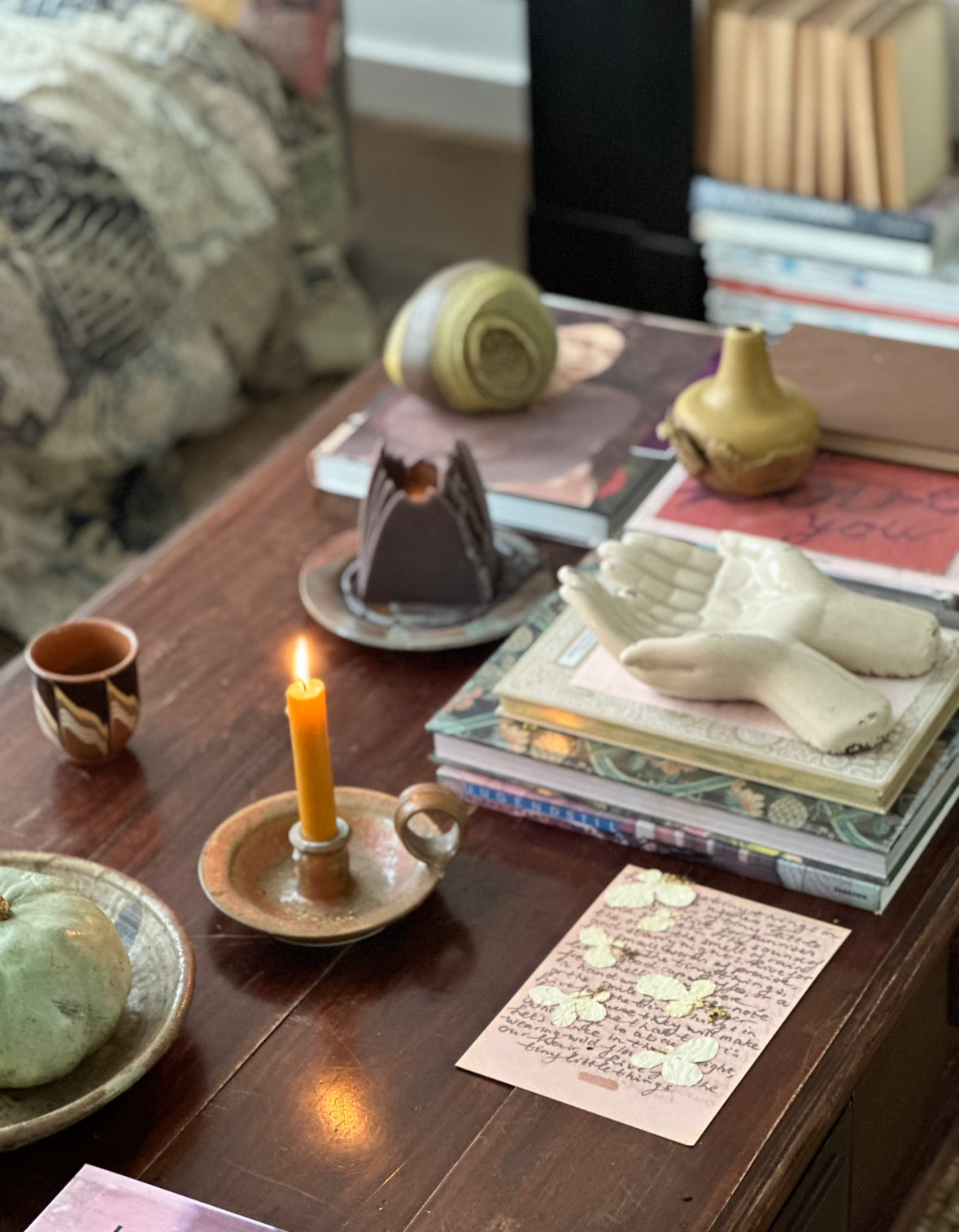 Handwritten pink note with cursive text beside a lit candle and ceramic hands sculpture on a cluttered wooden coffee table.