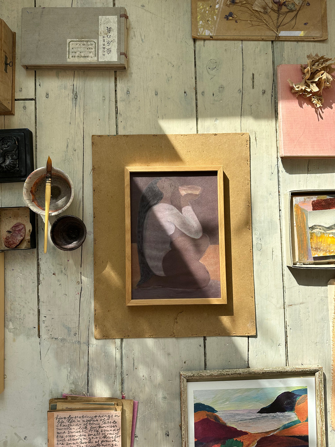 A framed artwork of a woman with long black hair, sitting cross-legged and holding a bowl, rendered in soft muted purple and brown tones, placed on a wooden surface with natural sunlight casting shadows.
