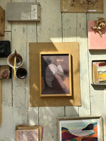 A framed artwork of a woman with long black hair, sitting cross-legged and holding a bowl, rendered in soft muted purple and brown tones, placed on a wooden surface with natural sunlight casting shadows.