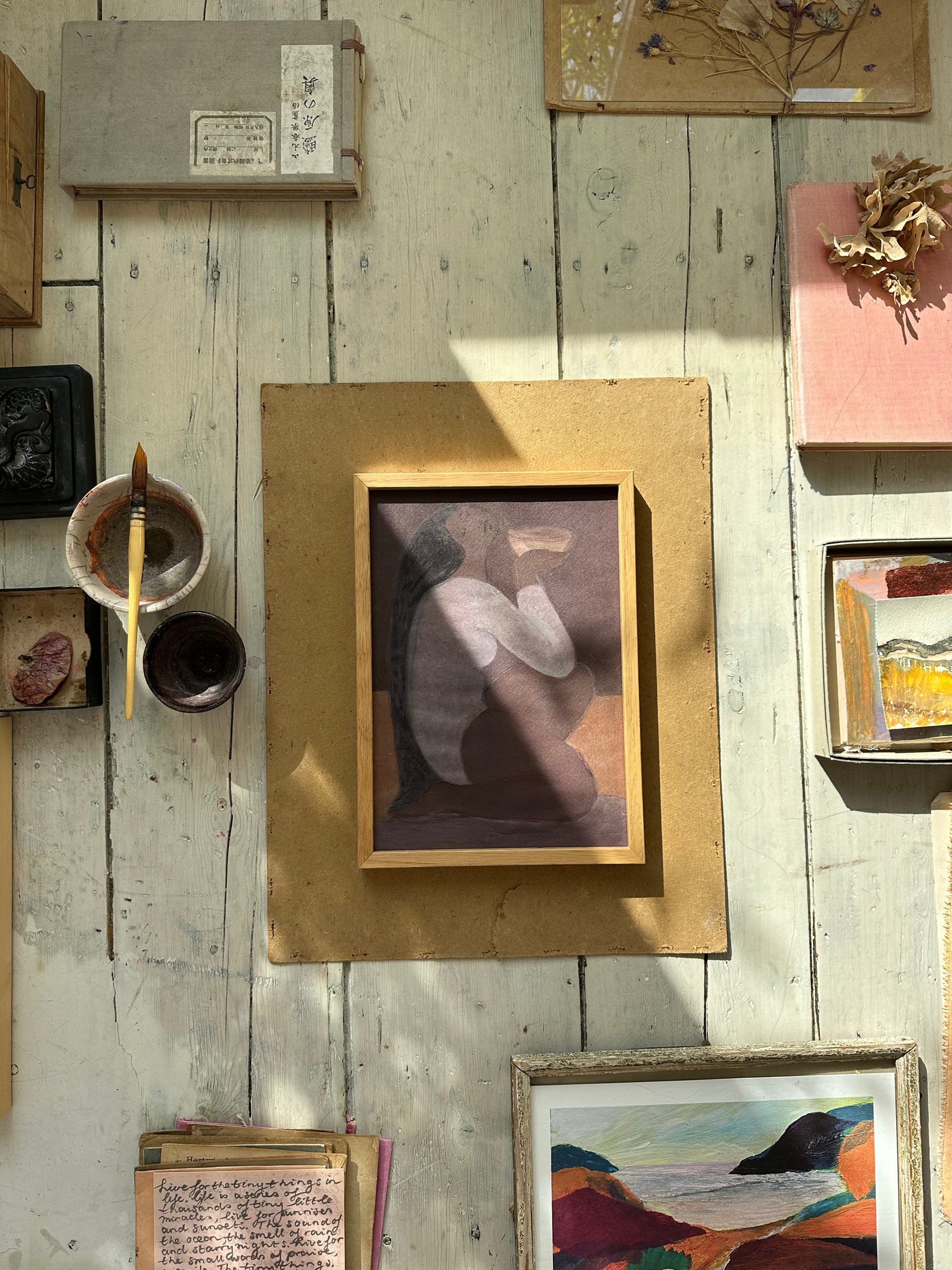 A framed artwork of a woman with long black hair, sitting cross-legged and holding a bowl, rendered in soft muted purple and brown tones, placed on a wooden surface with natural sunlight casting shadows.