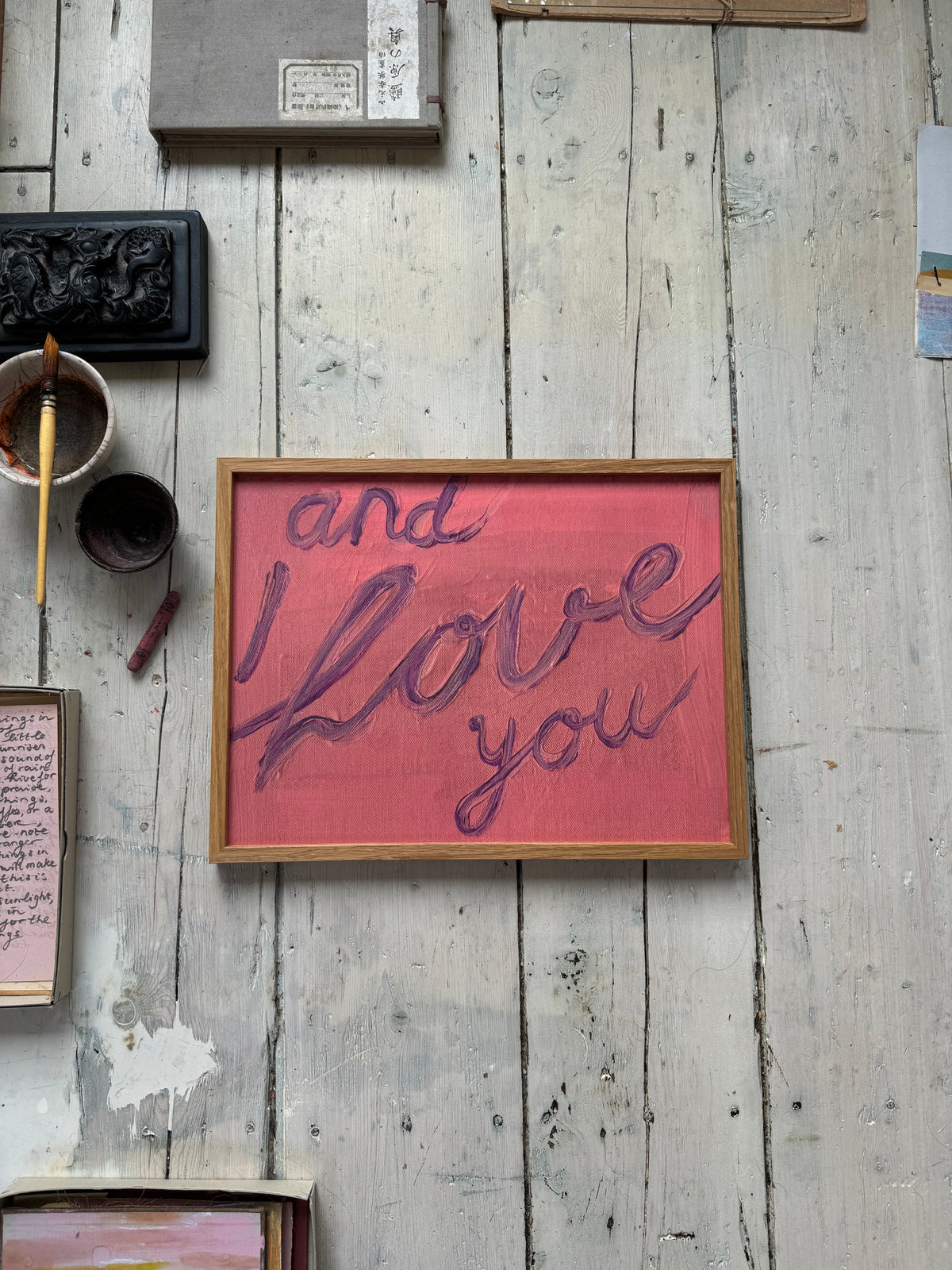 Framed artwork with handwritten text "and I love you" in purple on pink textured background, resting on rustic wooden floor.