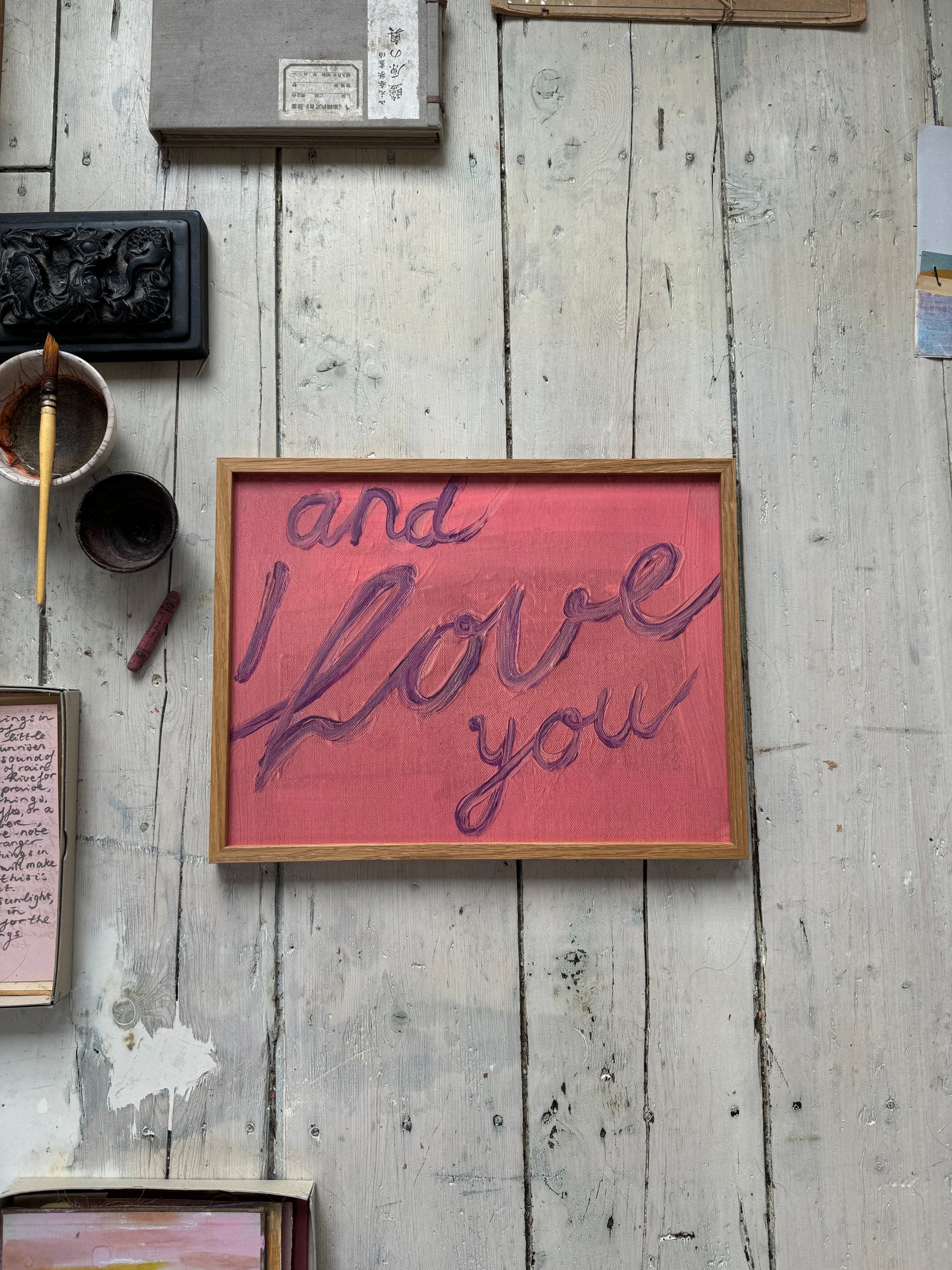 Framed artwork with handwritten text "and I love you" in purple on pink textured background, resting on rustic wooden floor.