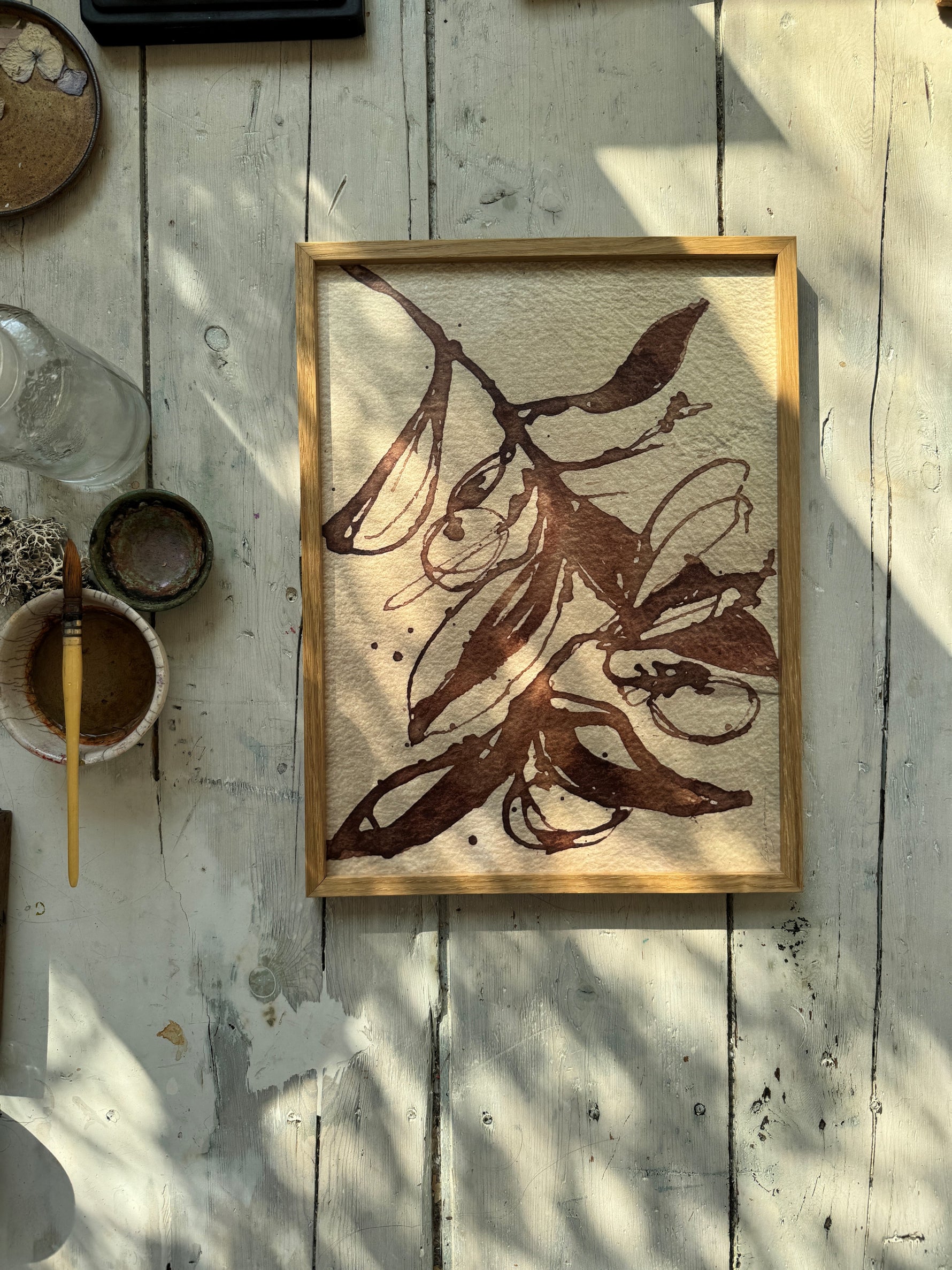 Framed brown botanical ink painting of leaves and seed pods on textured beige paper with natural light on wooden floor.