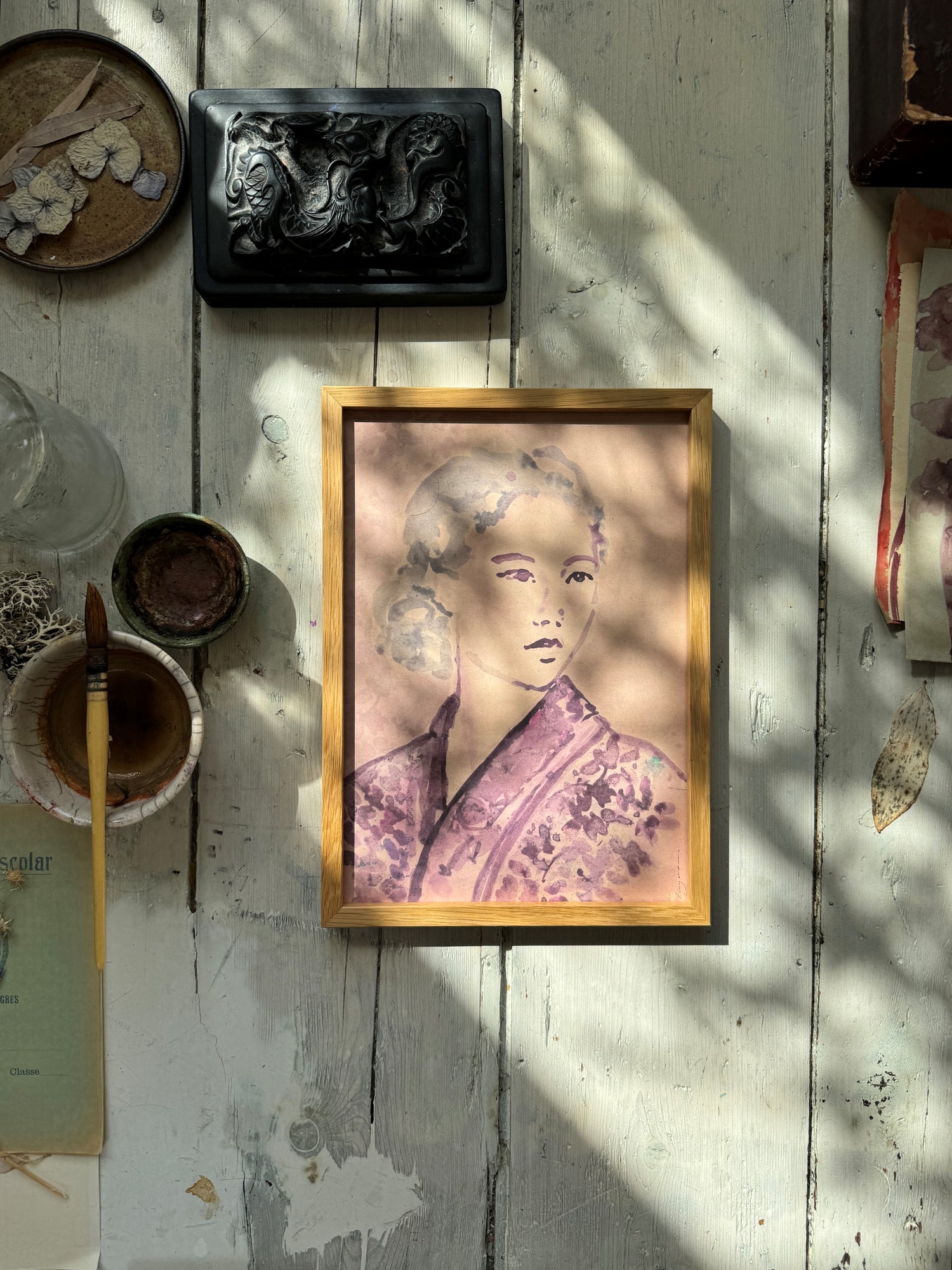 Artistic workspace with framed watercolor portrait of woman in kimono, black carved box, paintbrush, and bowls on wood floor.