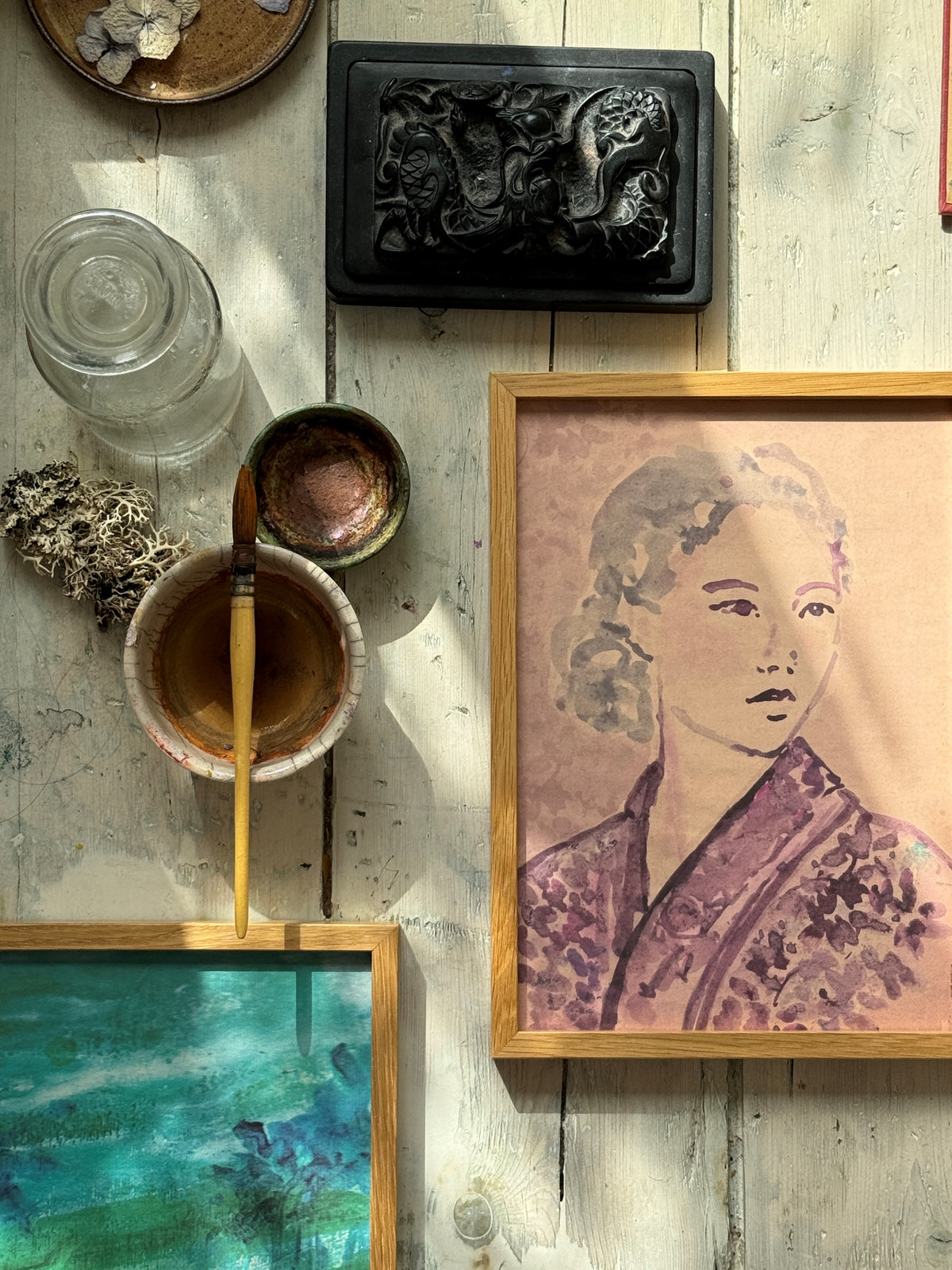 Close-up of framed kimono portrait and painting supplies on rustic wooden floor, natural light casting shadows.