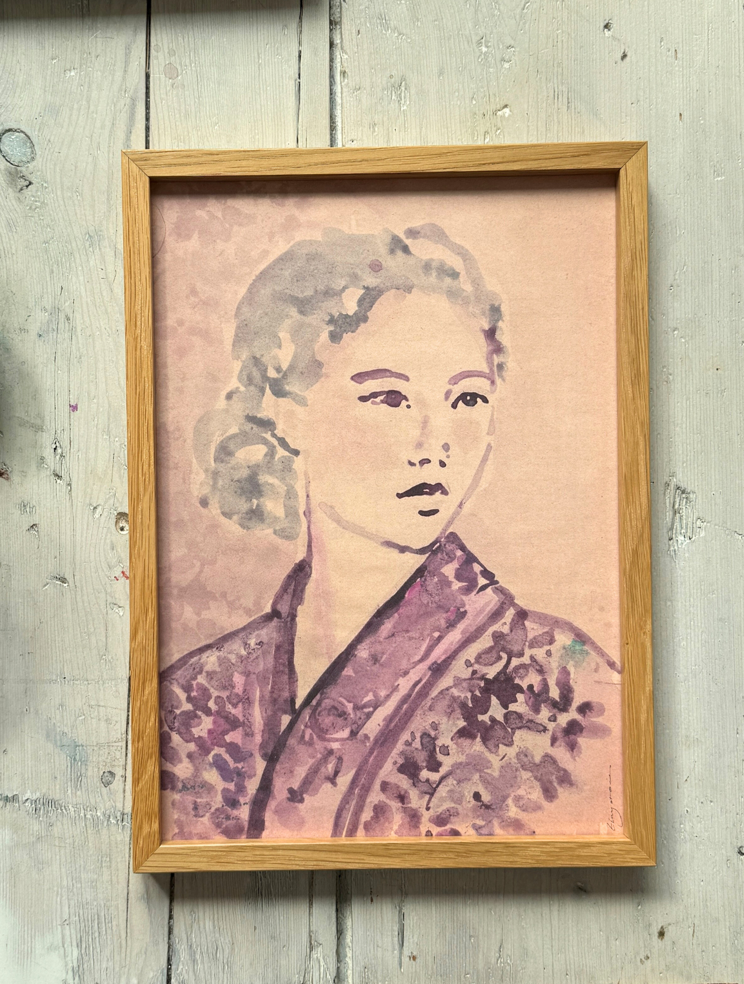 Framed watercolor portrait of woman in floral kimono with soft purple hues on rustic wooden floor with paintbrush nearby.