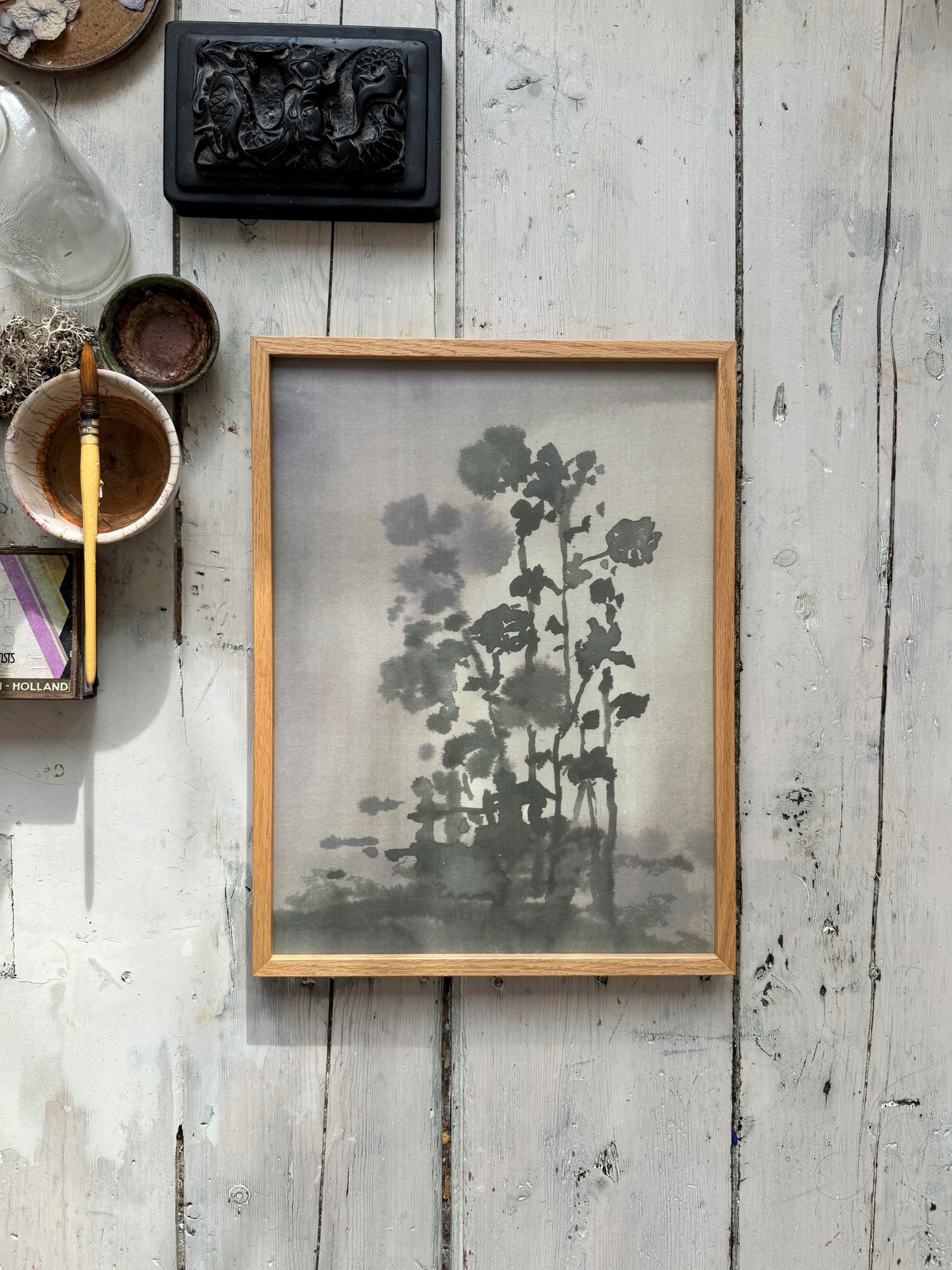 Artistic workspace with framed monochrome flower ink painting, paintbrush, bowls, and carved black box on wood floor.