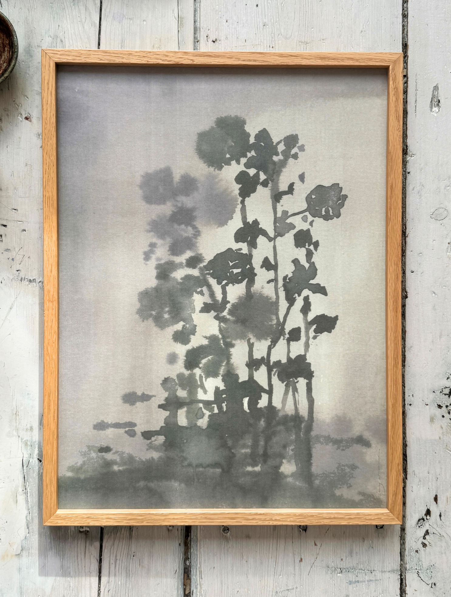 Framed monochrome ink painting of tall flowers and stems with soft shadows on rustic wooden floor.