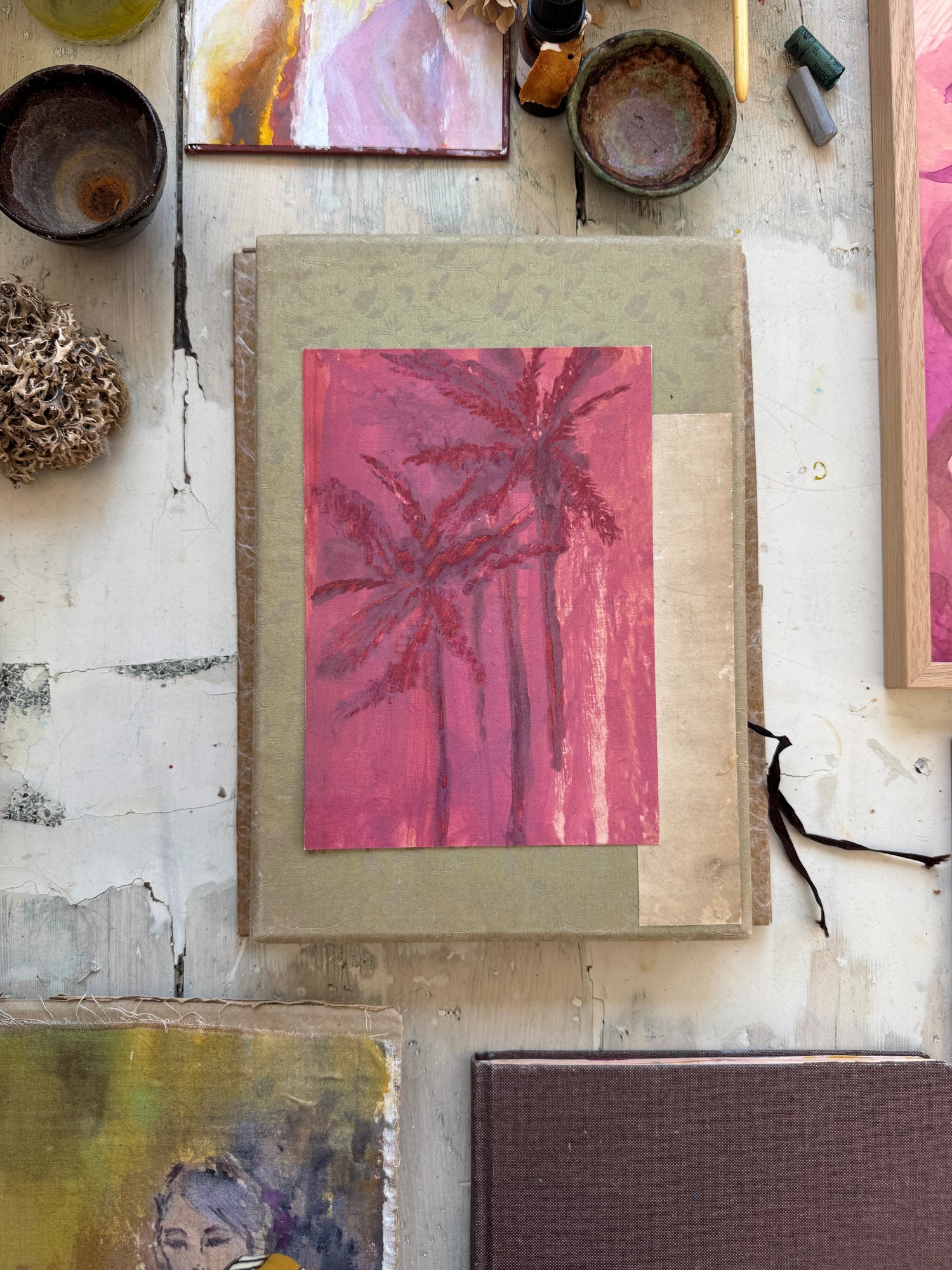 Pink palm tree watercolor painting on top of textured green book with paintbrush and art supplies on white wooden floor