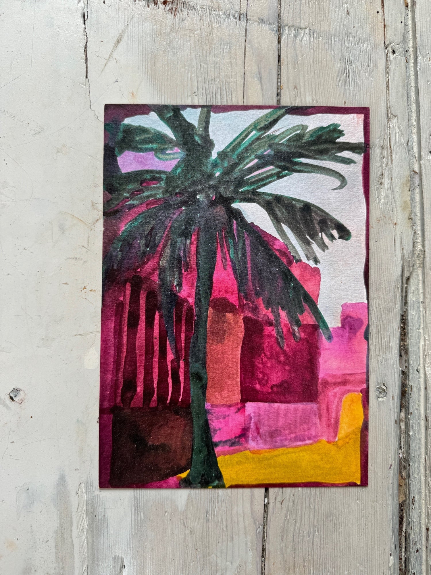 Abstract watercolor painting of a palm tree with dark green leaves against a vibrant pink and yellow background on wood floor.