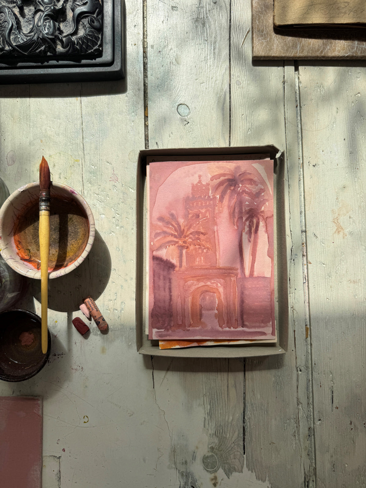Watercolor of an architectural archway with palm trees in pink and brown tones, beside paintbrush and pastel chalks
