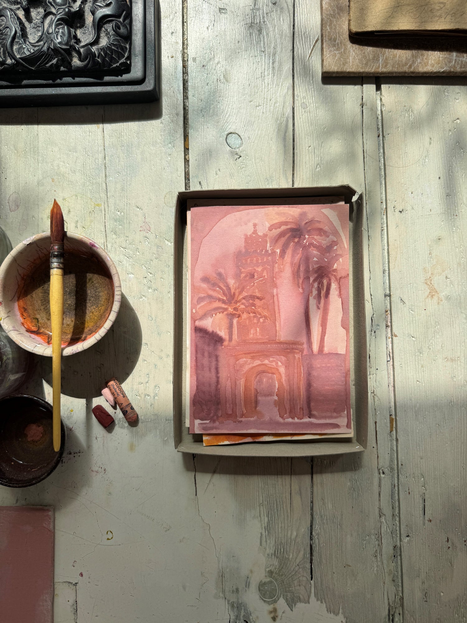 Watercolor of an architectural archway with palm trees in pink and brown tones, beside paintbrush and pastel chalks