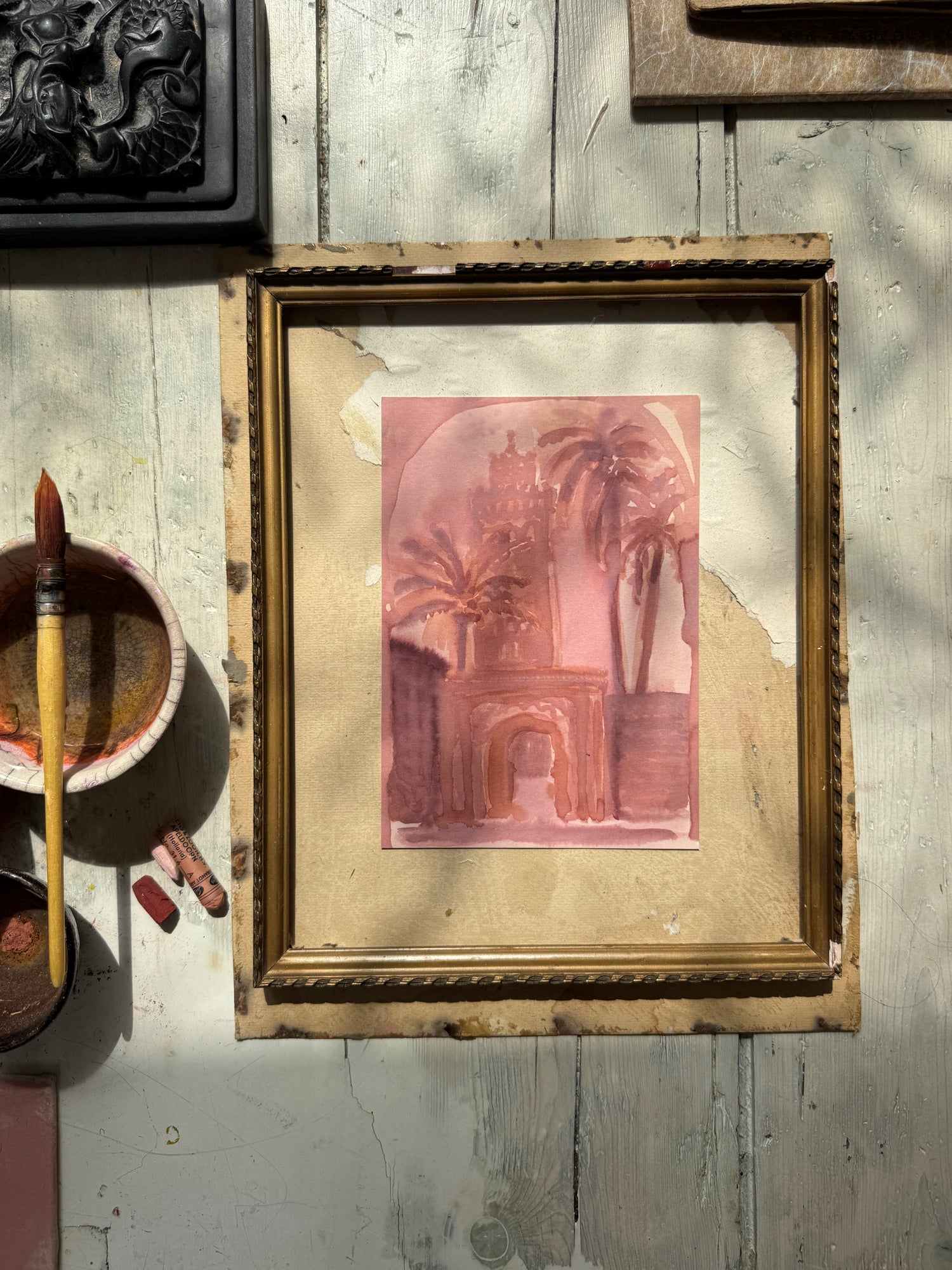 Watercolor of an architectural archway with palm trees in pink and brown tones, beside paintbrush and pastel chalks