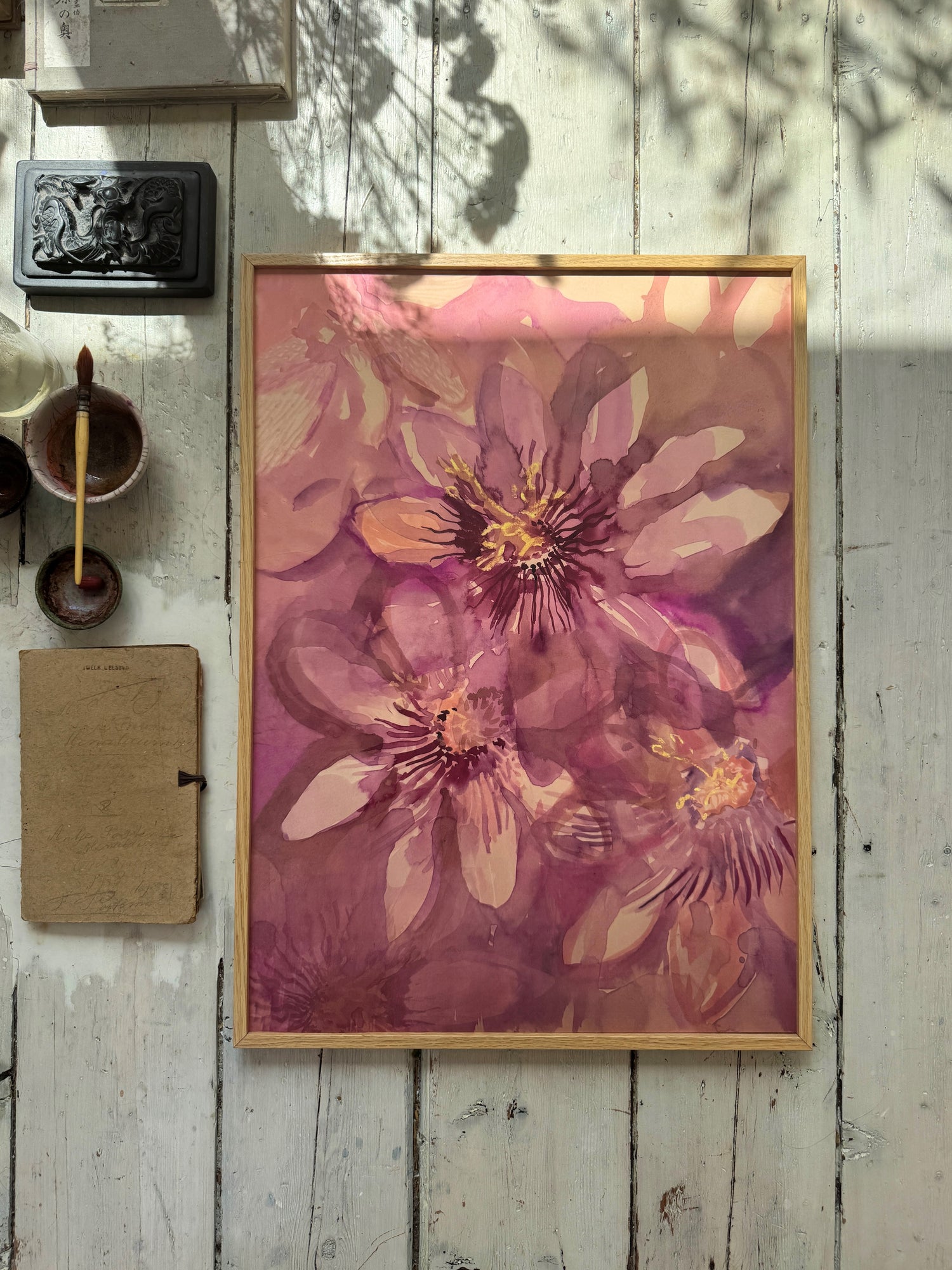 Framed watercolor painting of large pink flowers on rustic white wooden floor with paintbrush and paint cups beside it