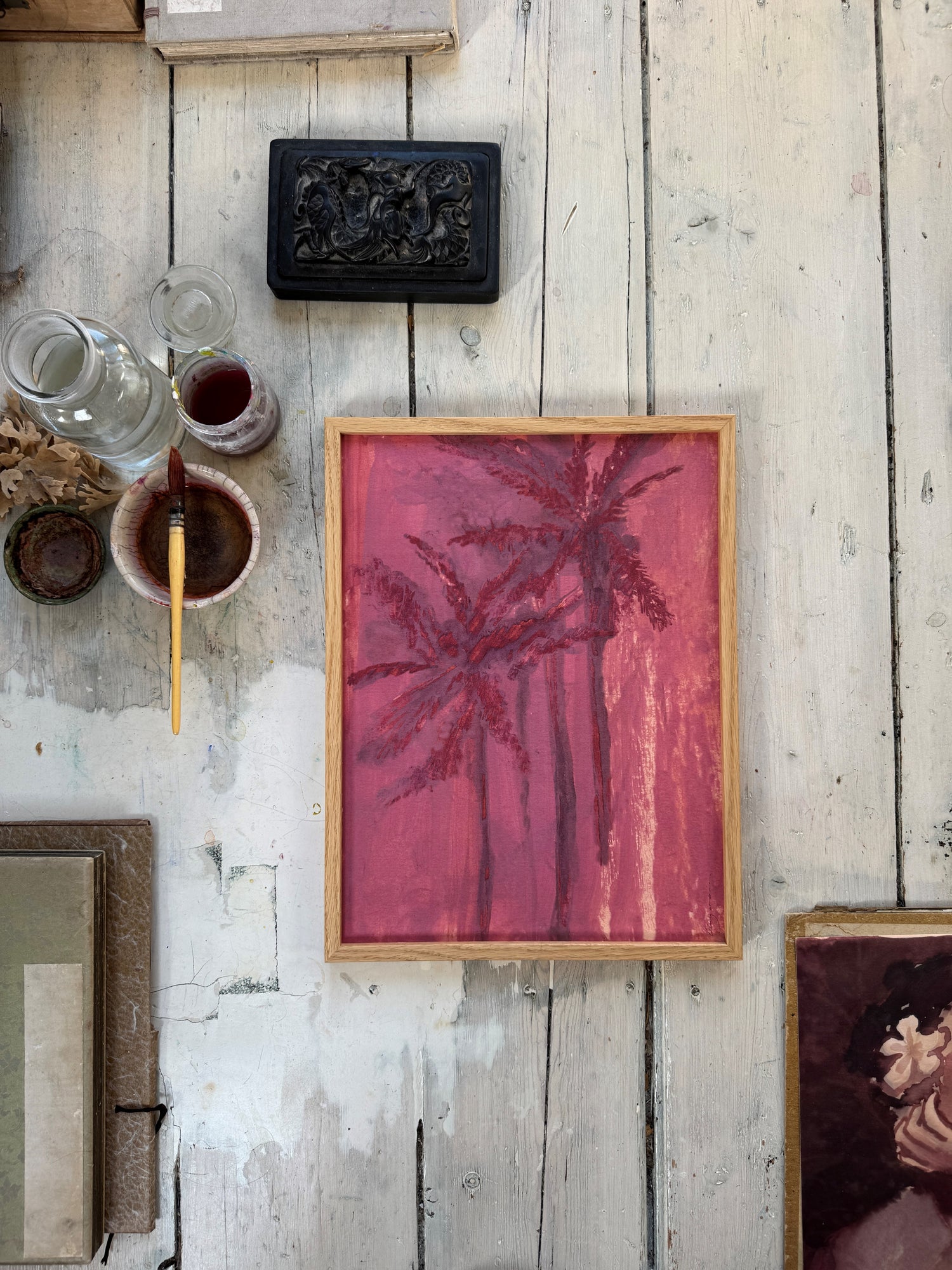 Framed watercolor painting of pink and purple palm trees on rustic white wooden floor beside paintbrush and glass bottle