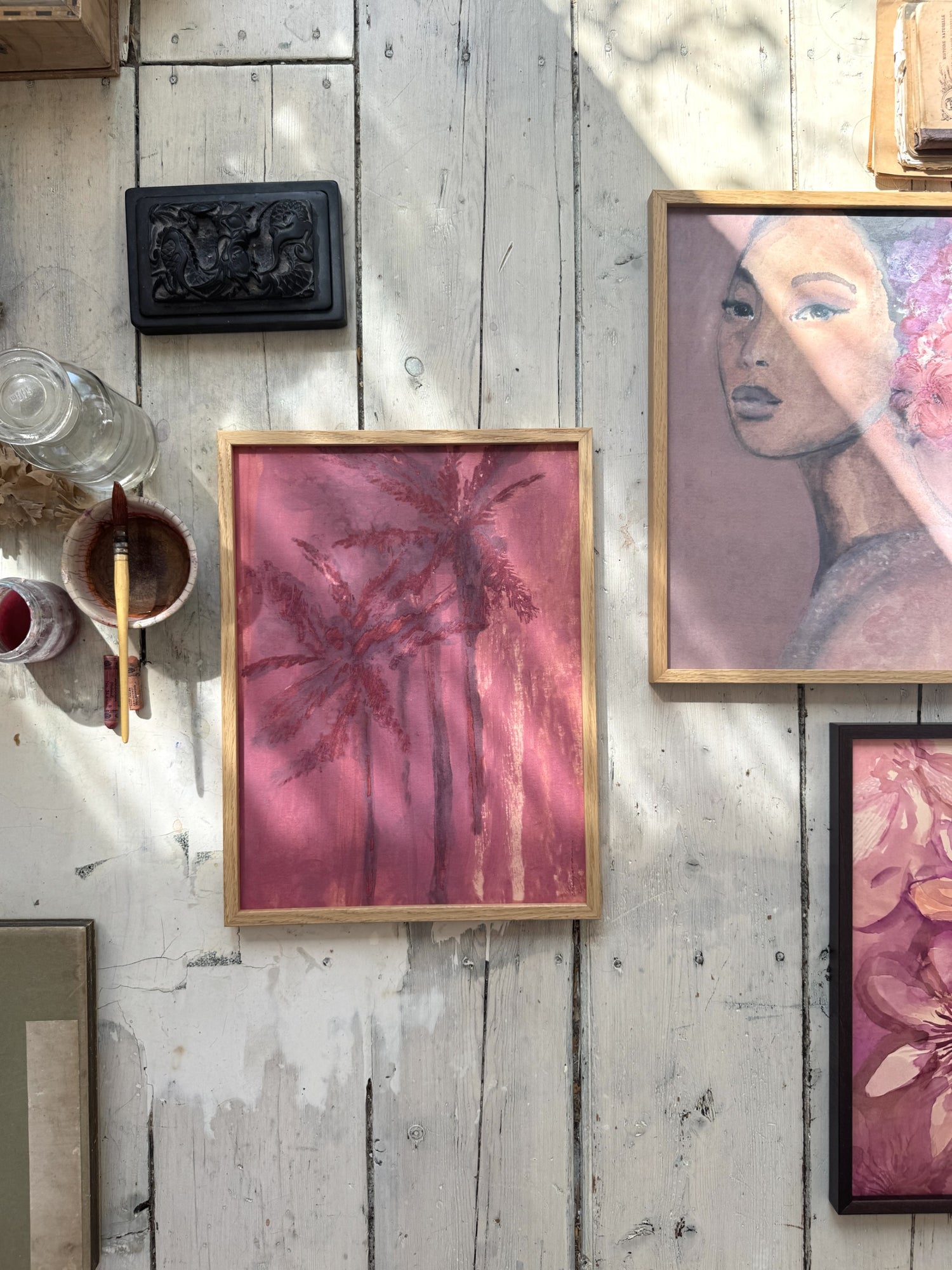 Close-up of framed palm tree painting with paintbrush, glass bottle, and paint cups on white wooden surface