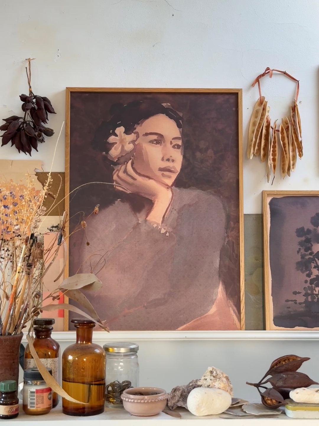 A pink-toned painting of lady with flower in hair against a white wall with glass bottles, shells and dried plants in front.