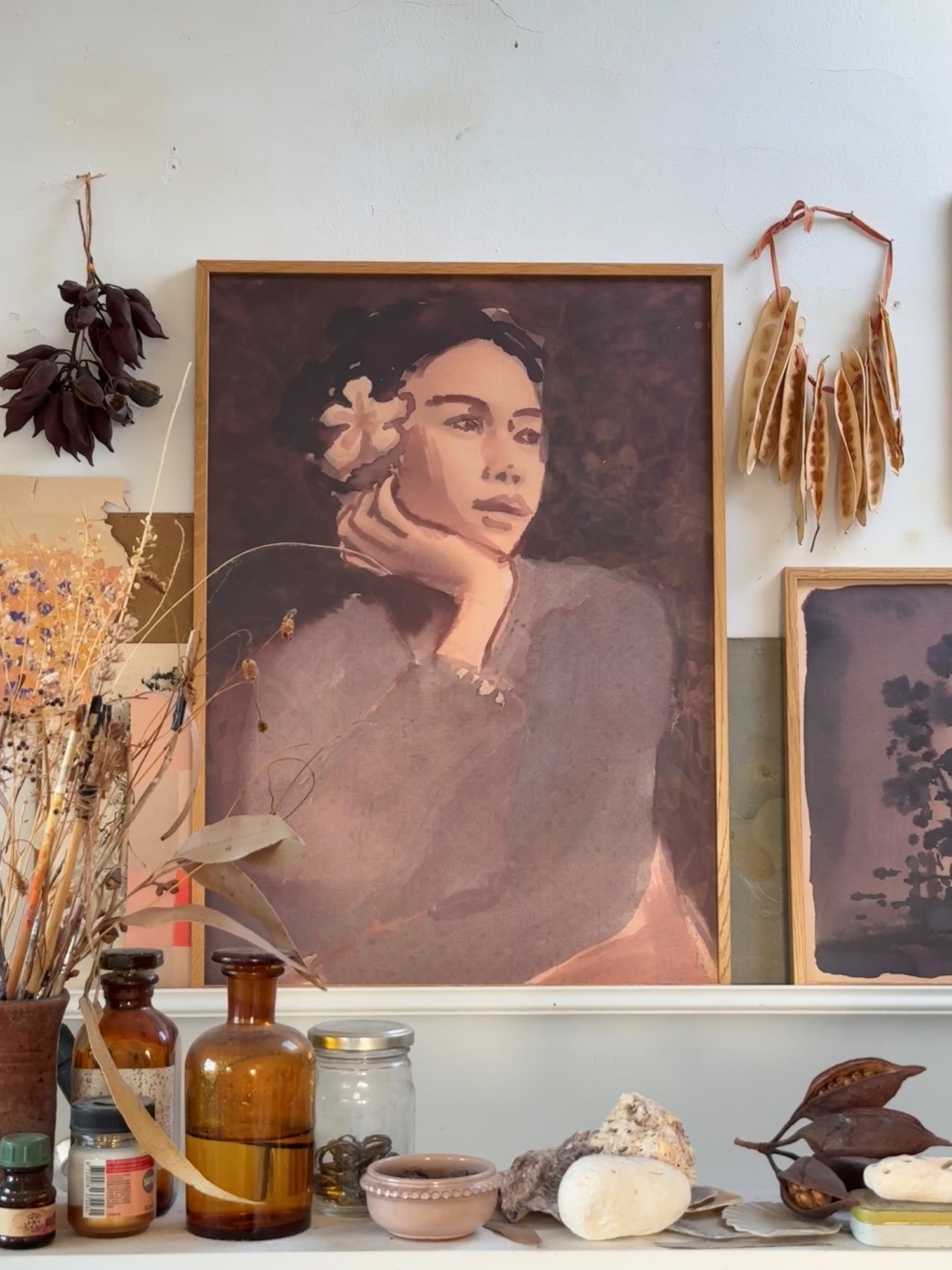 A pink-toned painting of lady with flower in hair against a white wall with glass bottles, shells and dried plants in front.