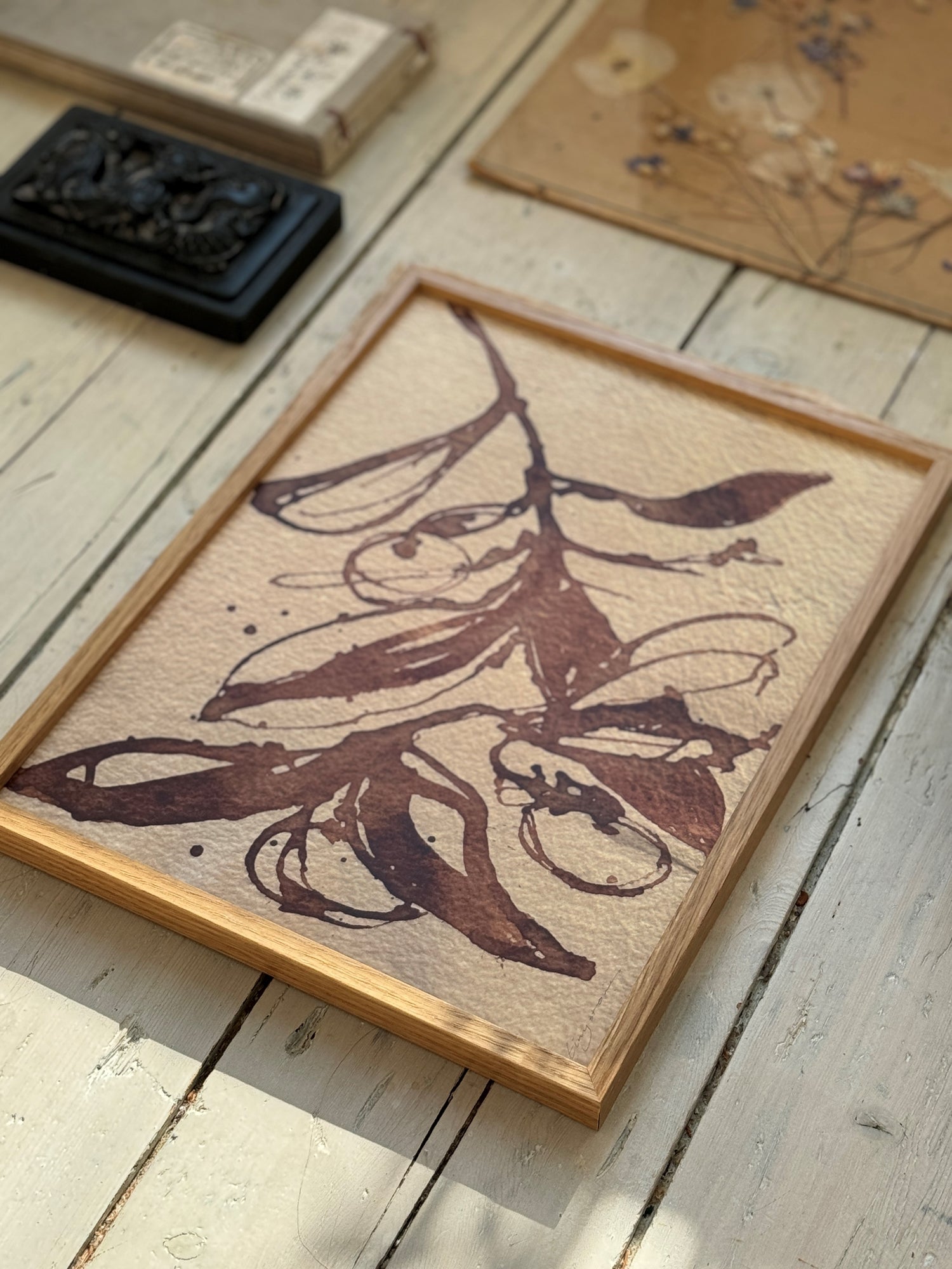 Angled view of framed botanical ink art depicting leaves and pods on beige textured paper with art supplies around.