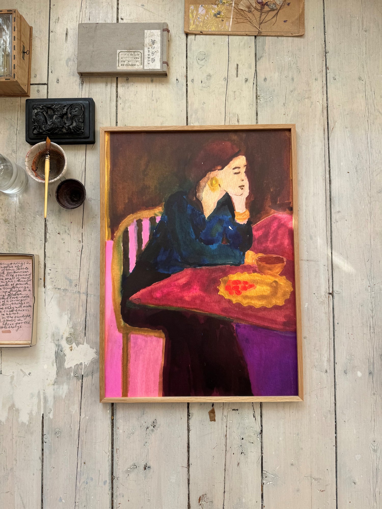 Framed vibrant figurative painting of a woman seated at a pink table on rustic wooden floor