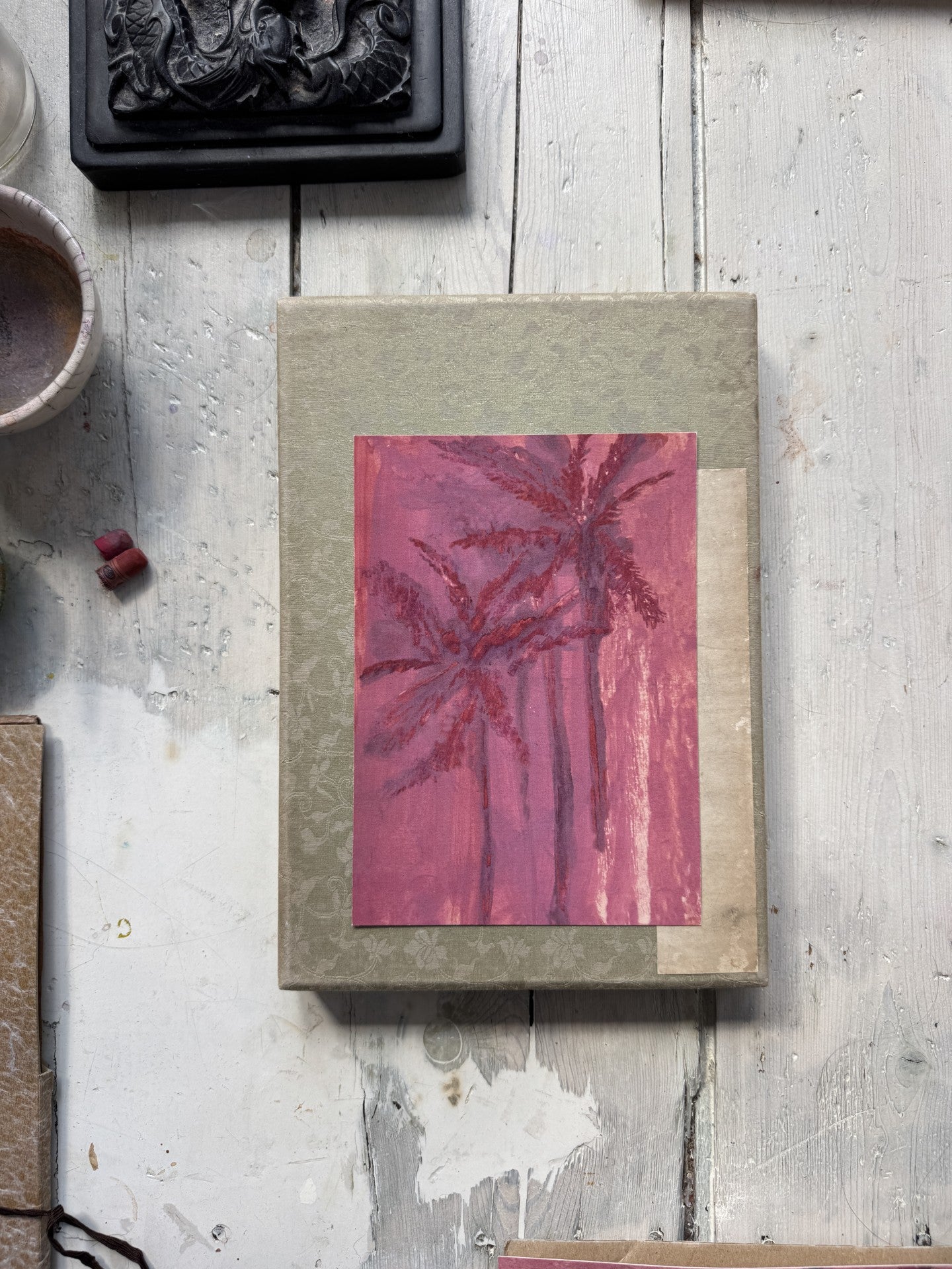 Top view of pink palm tree painting on textured green book with paintbrush, pastel chalks, and black carved object nearby
