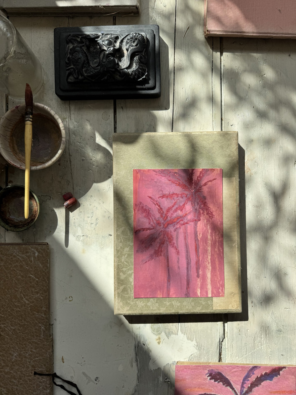 Pink palm tree watercolor painting on green book with sunlight and shadow on rustic white wooden surface and paintbrush nearby