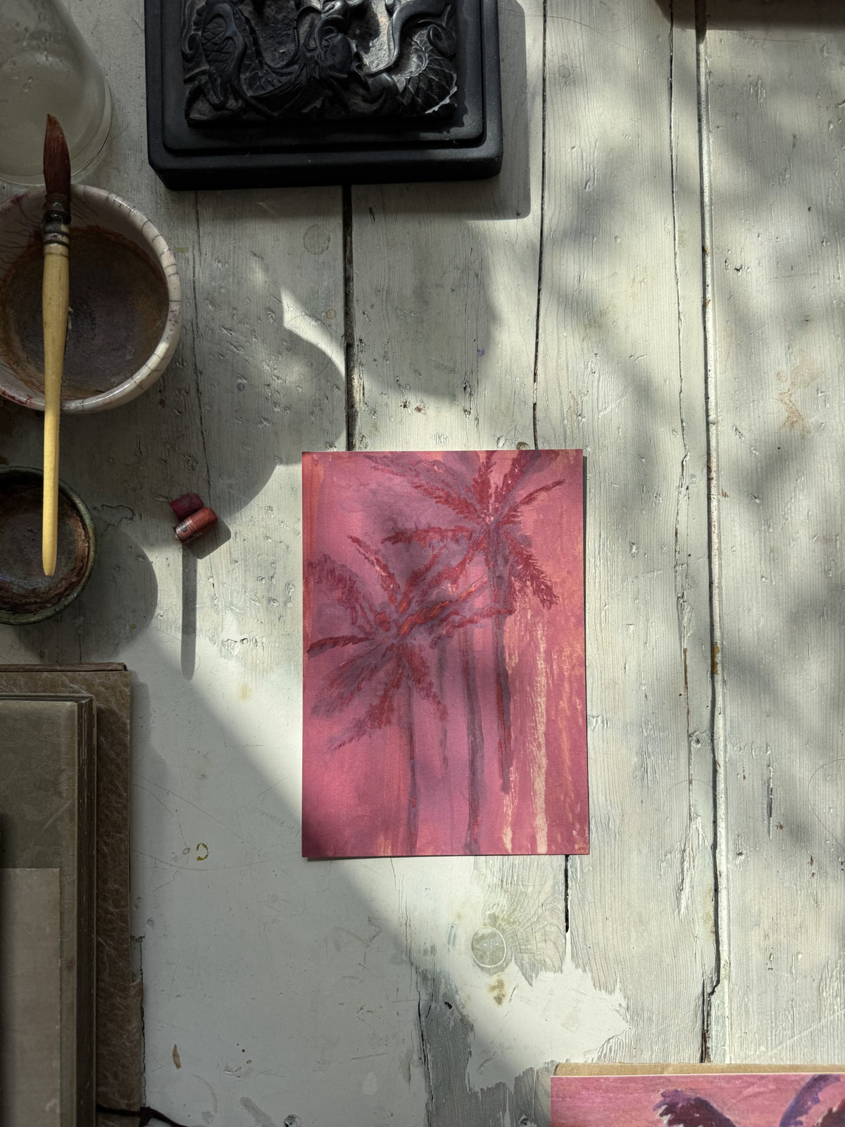 Pink palm tree painting placed on rustic white wooden surface with partial sunlight and art materials around it