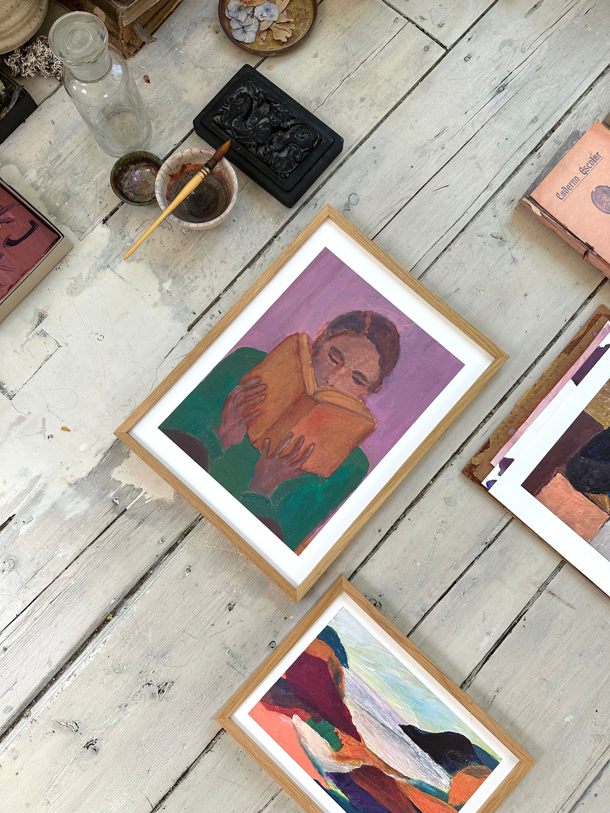 Angled close-up of framed reading figure painting surrounded by other colorful artworks on wooden floor.