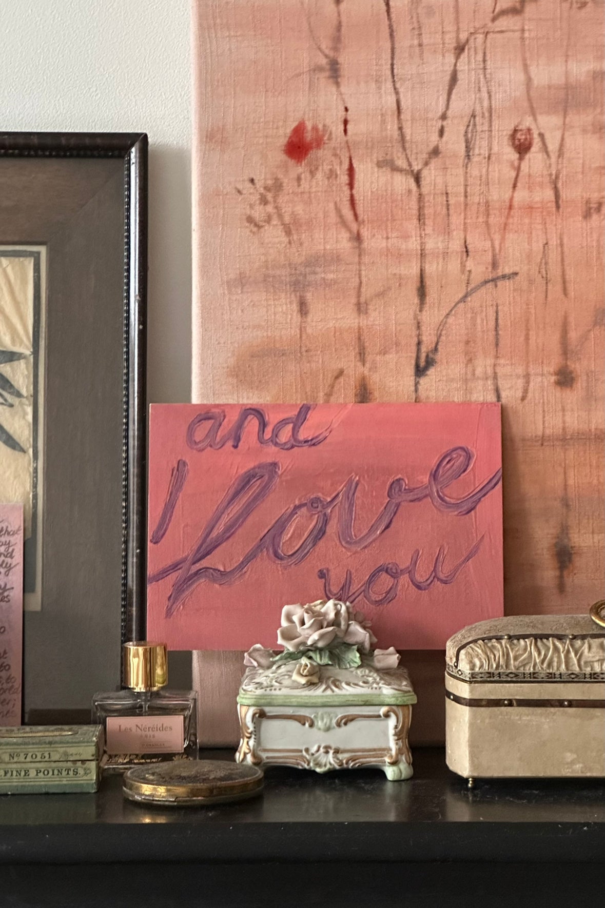 Artwork with handwritten text "and I love you" in purple on pink textured background, resting on wooden table with perfume in front.