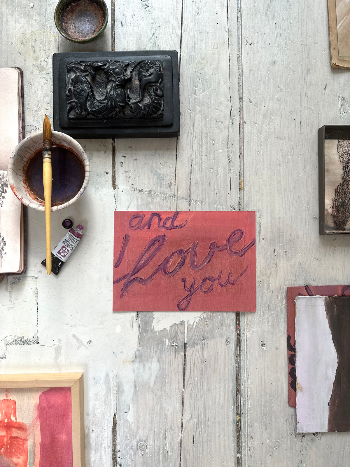 Artwork with handwritten text "and I love you" in purple on pink textured background, resting on rustic wooden floor with paint brush nearby.