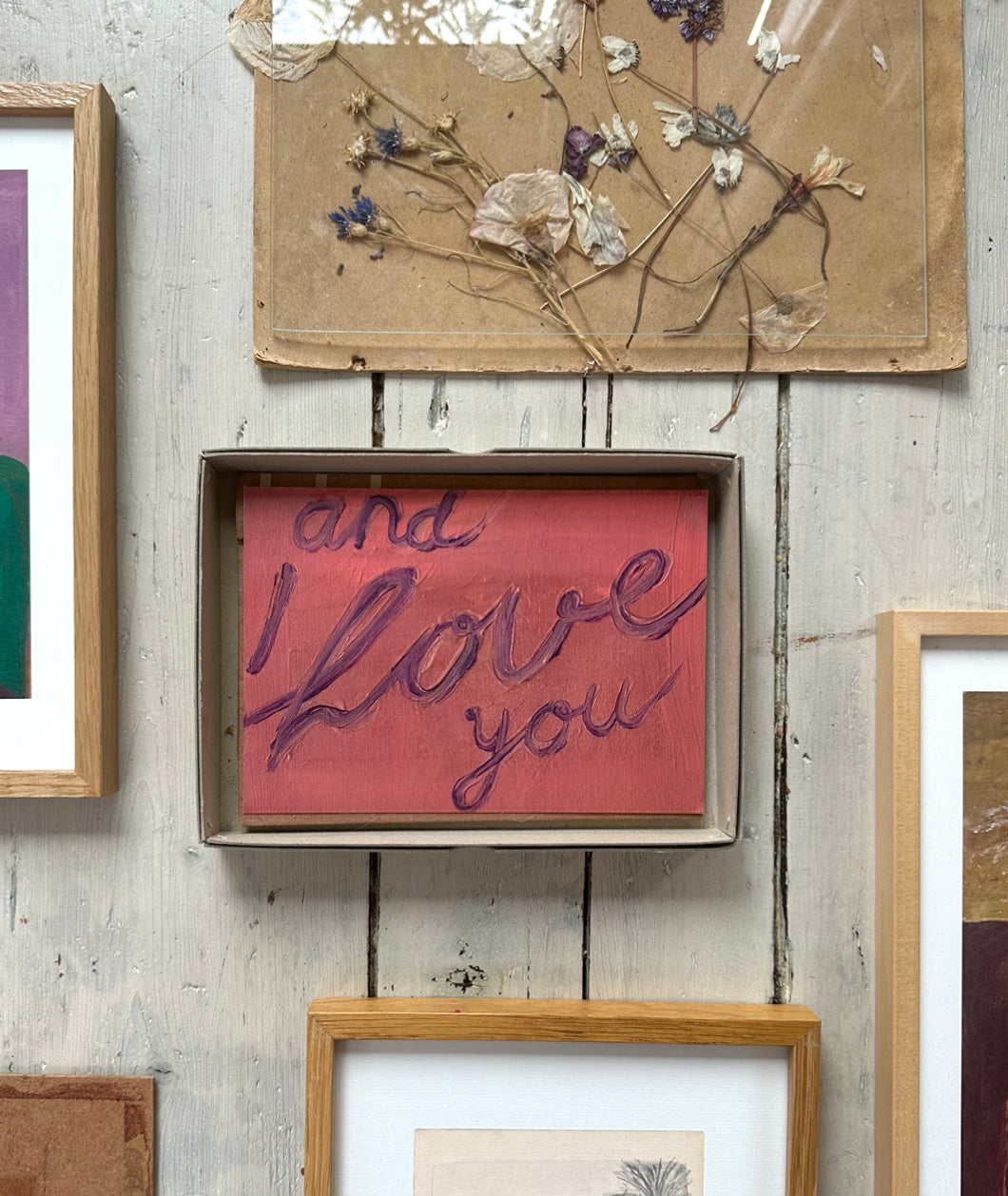Artwork with handwritten text "and I love you" in purple on pink textured background, resting on rustic wooden floor.