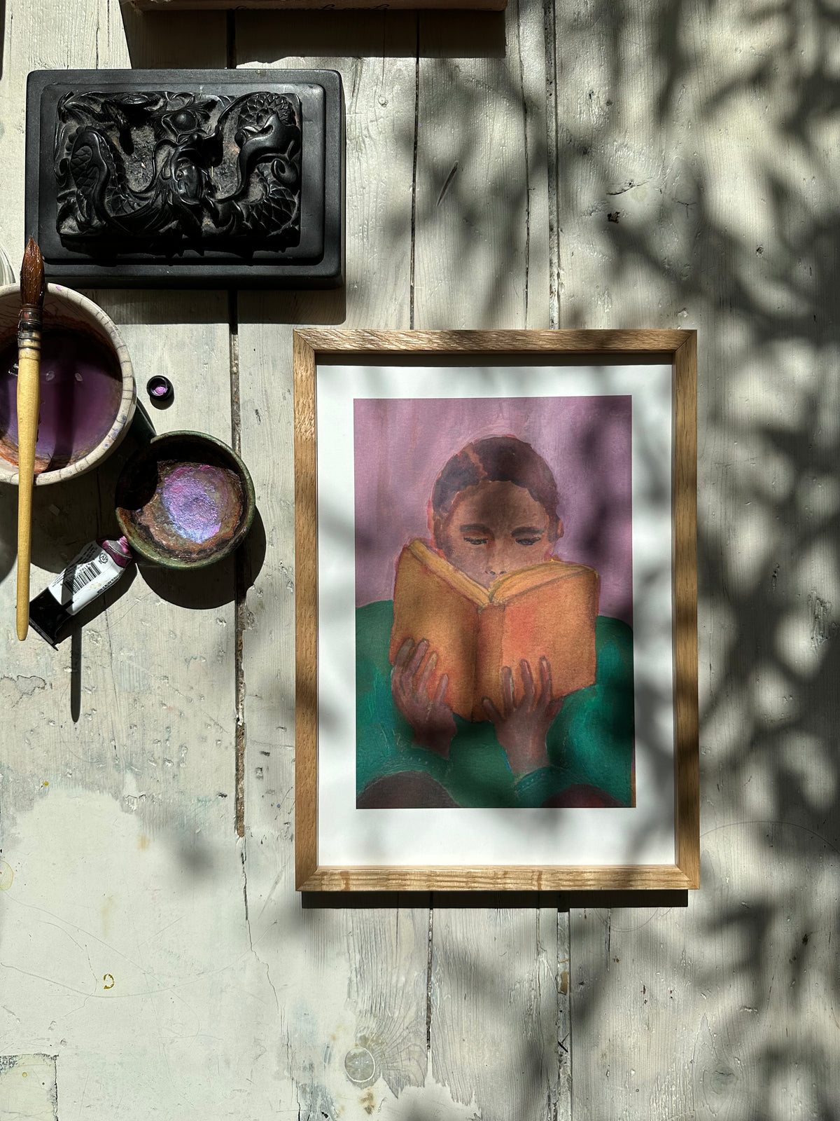 Purple-themed painting of person reading book displayed on white rustic floor wih art supplies nearby.