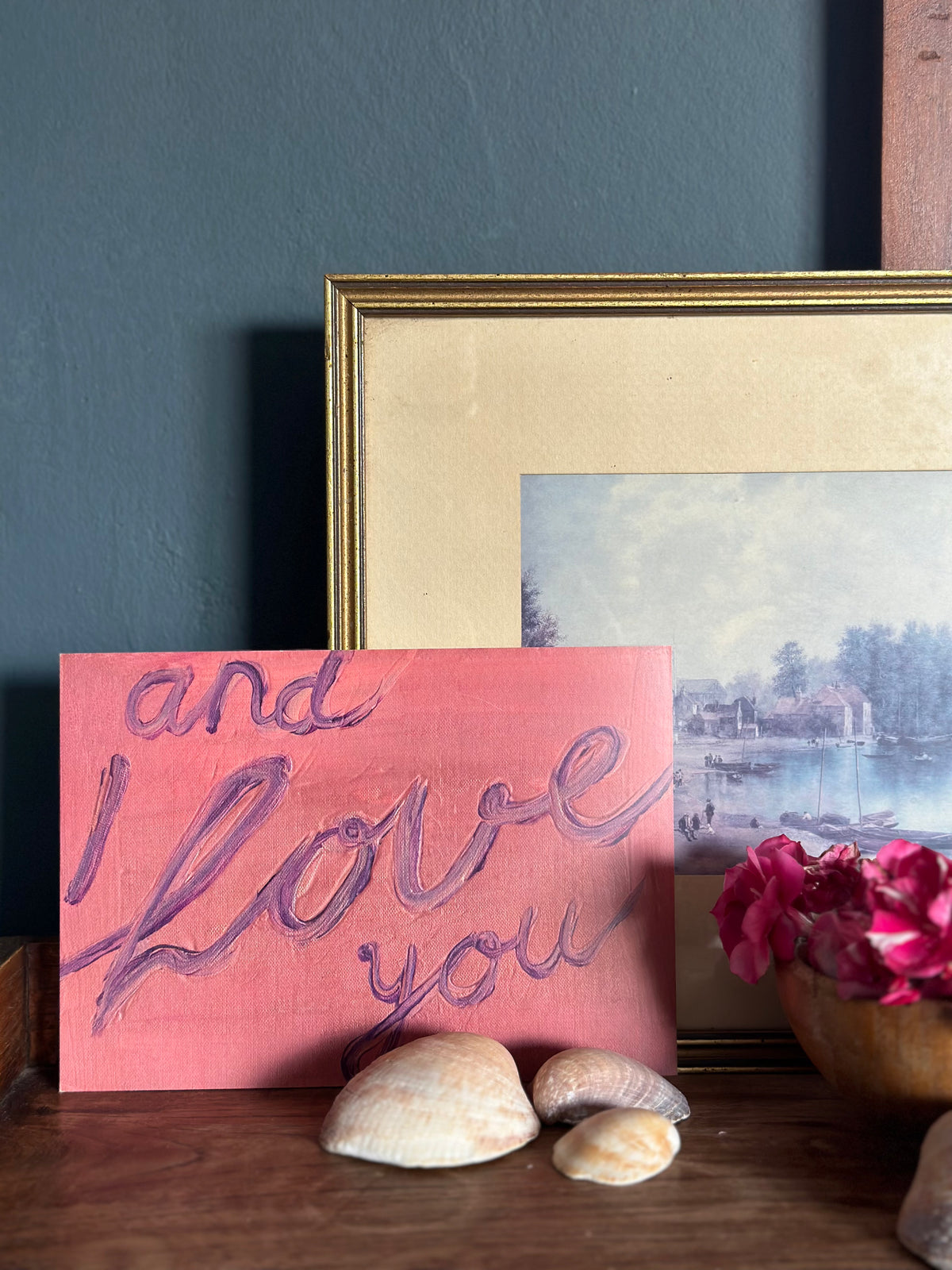 Artwork with handwritten text "and I love you" in purple on pink textured background, resting on rustic wooden table with shells in front.