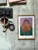 Framed painting of person reading orange book, purple background, on rustic wooden floor with paintbrush and art supplies nearby.