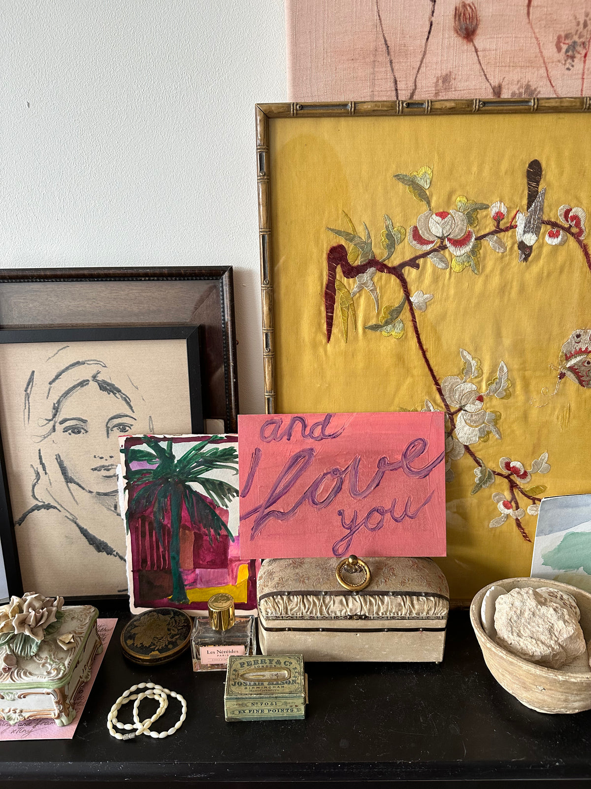 Artwork with handwritten text "and I love you" in purple on pink textured background, resting on wooden table with other paintings around.