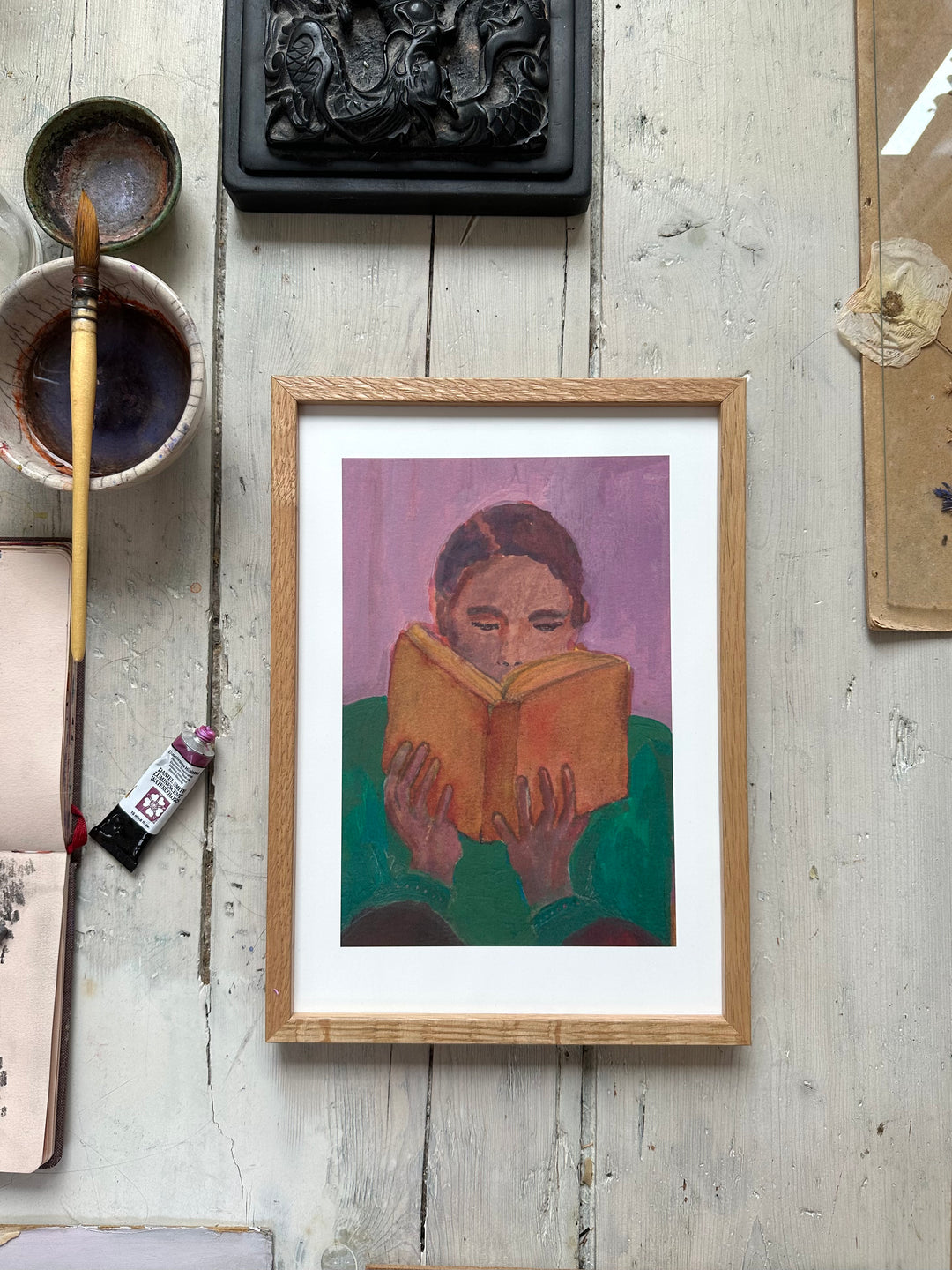 Framed painting of person reading orange book, purple background, on rustic wooden floor with paintbrush and art supplies nearby.