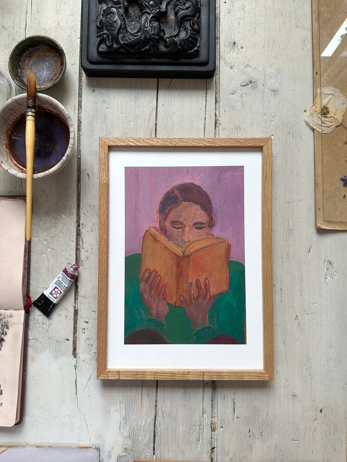 Framed painting of person reading orange book, purple background, on rustic wooden floor with paintbrush and art supplies nearby.
