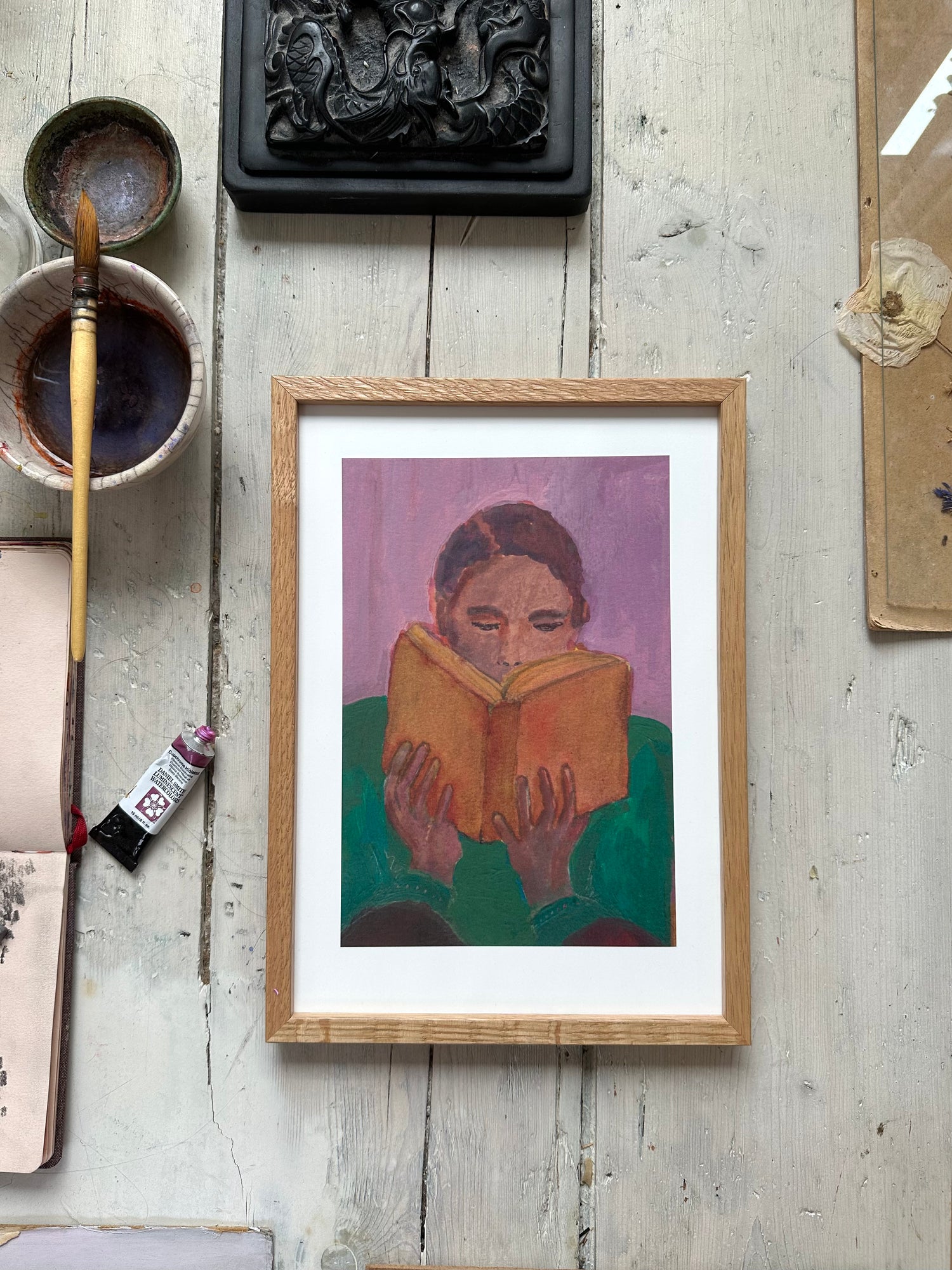 Framed painting of person reading orange book, purple background, on rustic wooden floor with paintbrush and art supplies nearby.