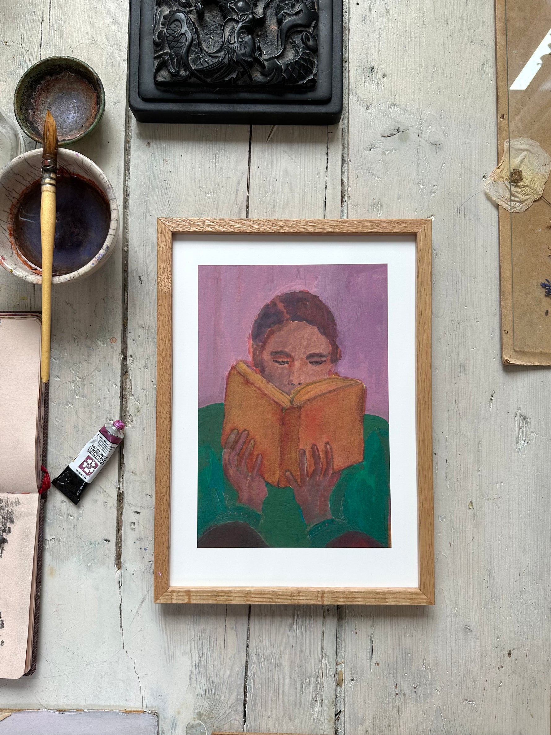 Framed painting of person reading orange book, purple background, on rustic wooden floor with paintbrush and art supplies nearby.