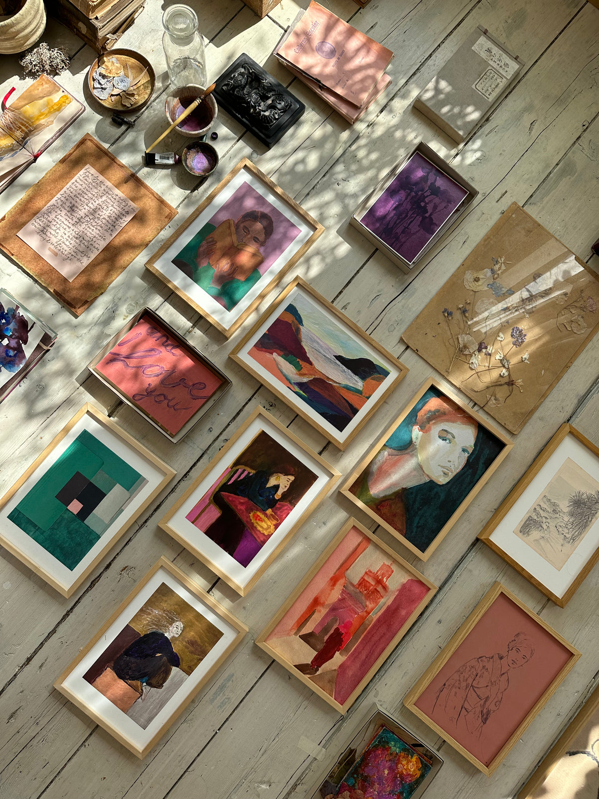 Overhead view of assorted framed paintings laid out on rustic wooden floor with natural sunlight highlighting textures.
