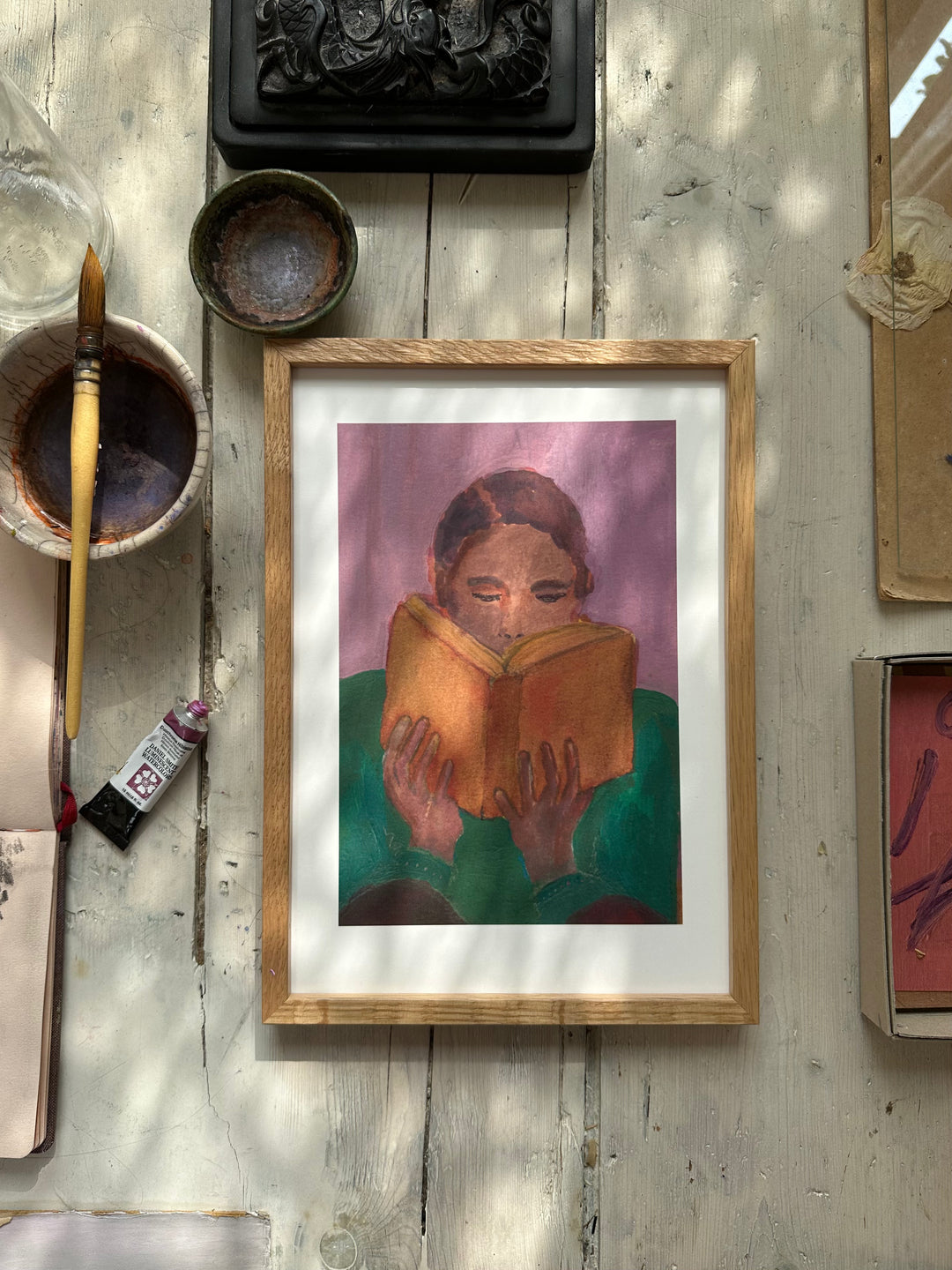 Purple-themed painting of person reading book displayed on white wall among other framed artworks and floral sketches.