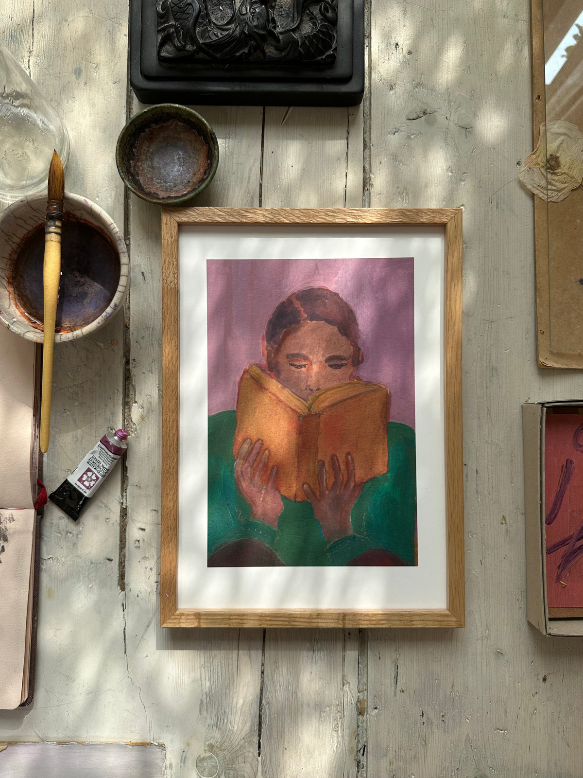 Purple-themed painting of person reading book displayed on white wall among other framed artworks and floral sketches.