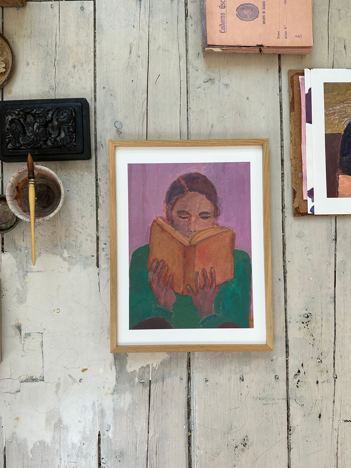 Purple-themed painting of person reading book displayed on white wall among other framed artworks.