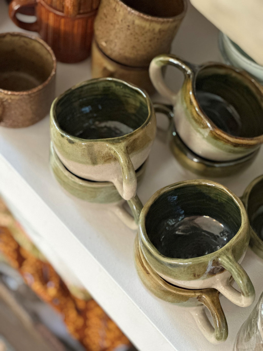 Stack of handmade ceramic cups with green glaze and beige base on white shelf with warm natural light.