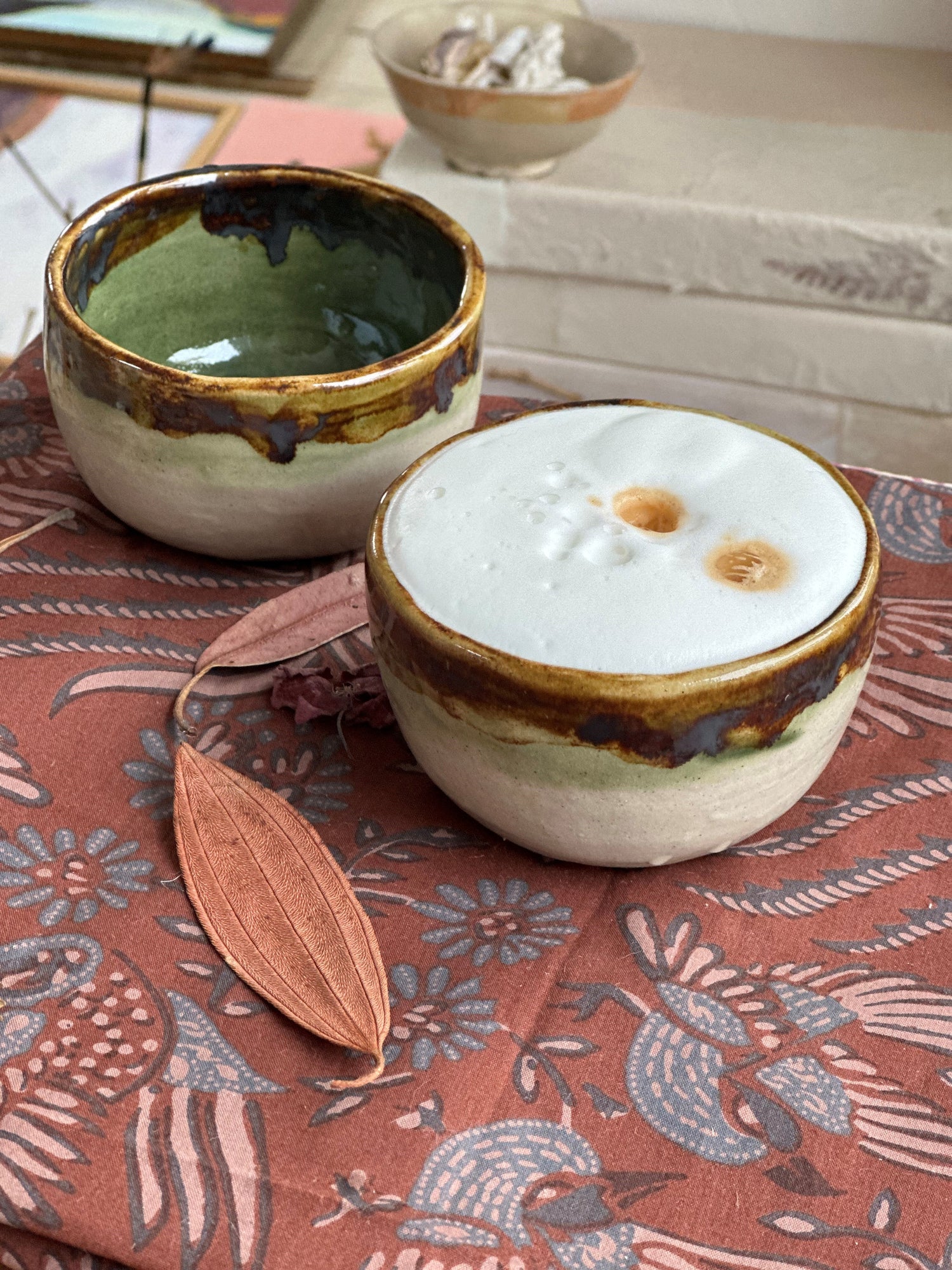 Close-up of ceramic cup filled with frothy coffee on patterned cloth with dried leaves beside it.