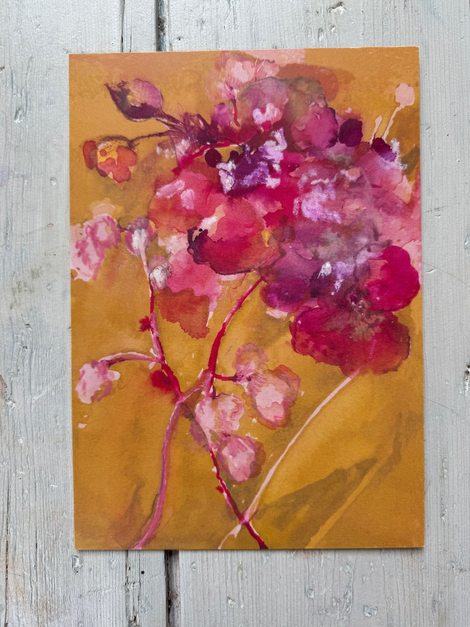 Watercolor painting of pink and purple flowers on warm ochre background, displayed on rustic wooden floor.
