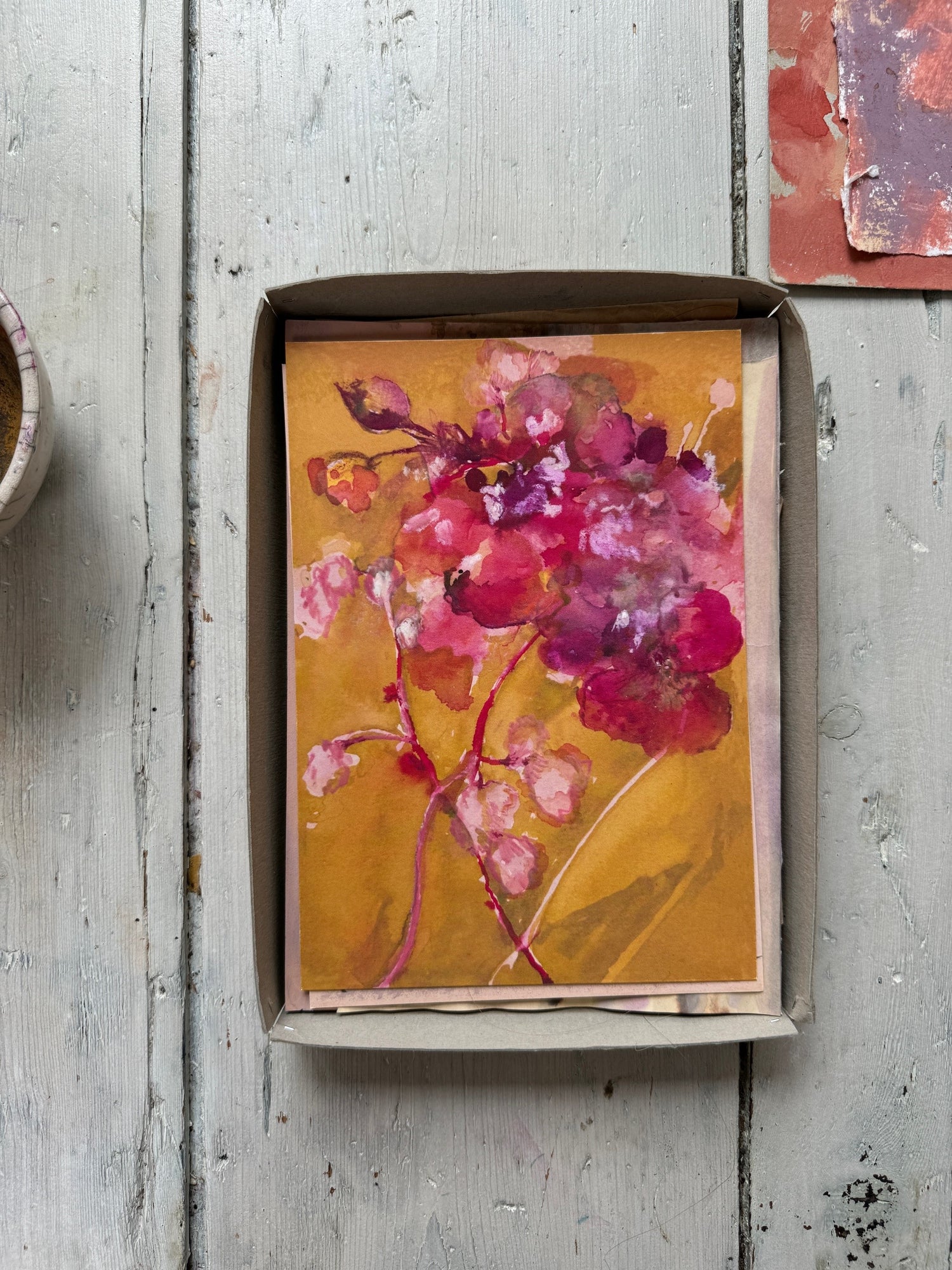 Watercolor painting of pink and purple flowers on warm ochre background, displayed on rustic wooden floor.