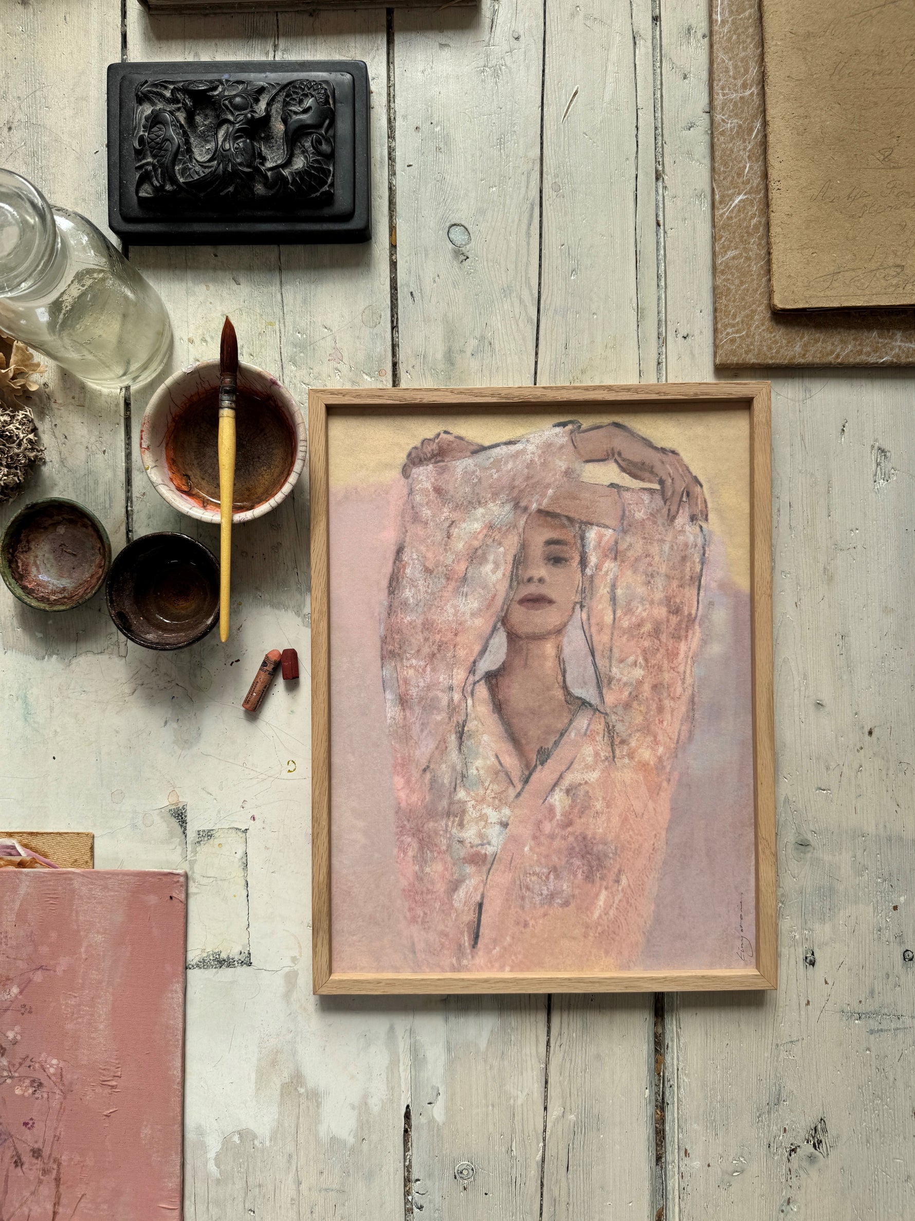 Framed watercolor portrait of a woman in sheer floral dress with arms raised surrounded by paintbrush and art supplies on wooden table
